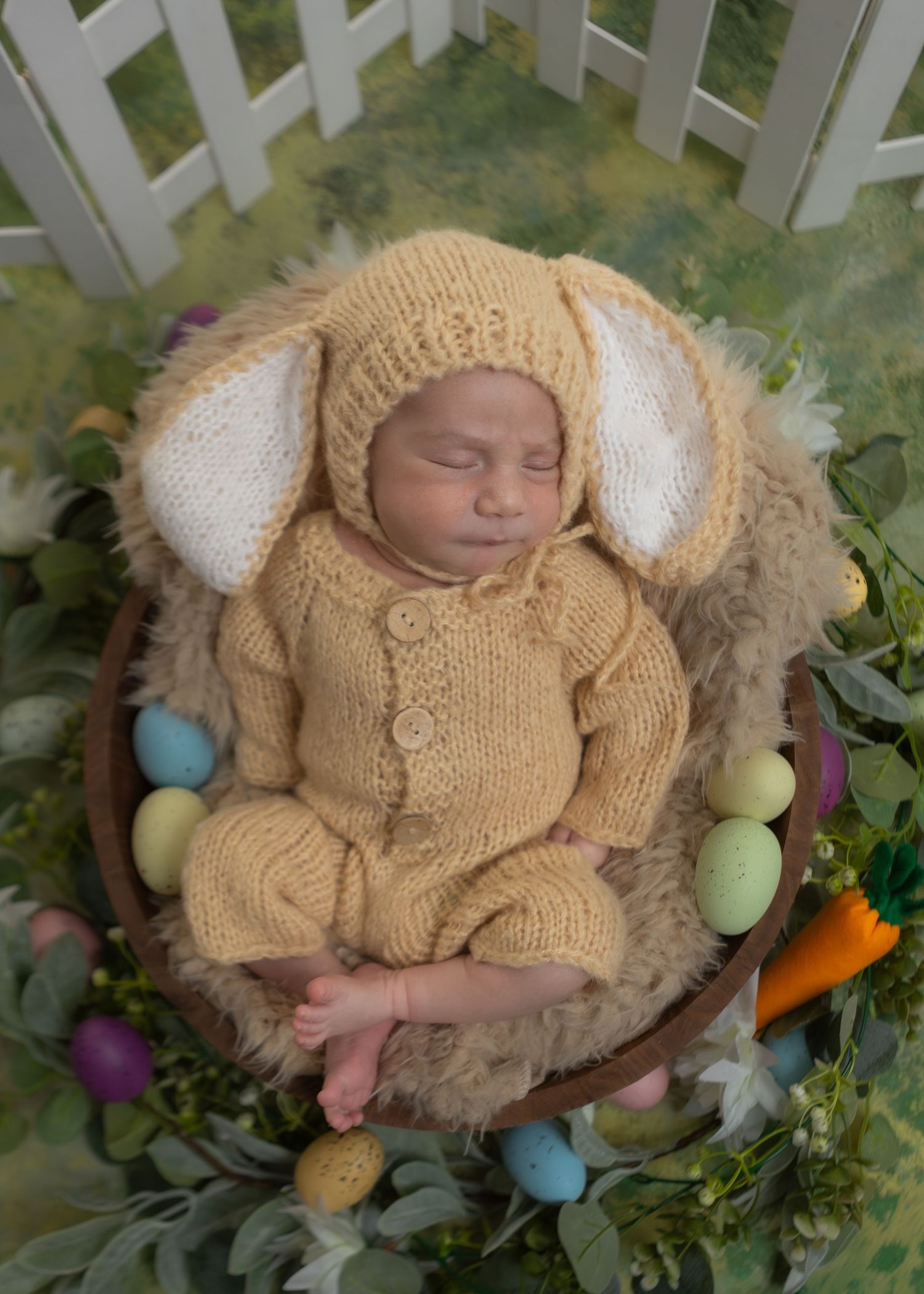 Sleeping newborn in knit bunny Easter outfit posed in wooden bowl with colorful eggs.