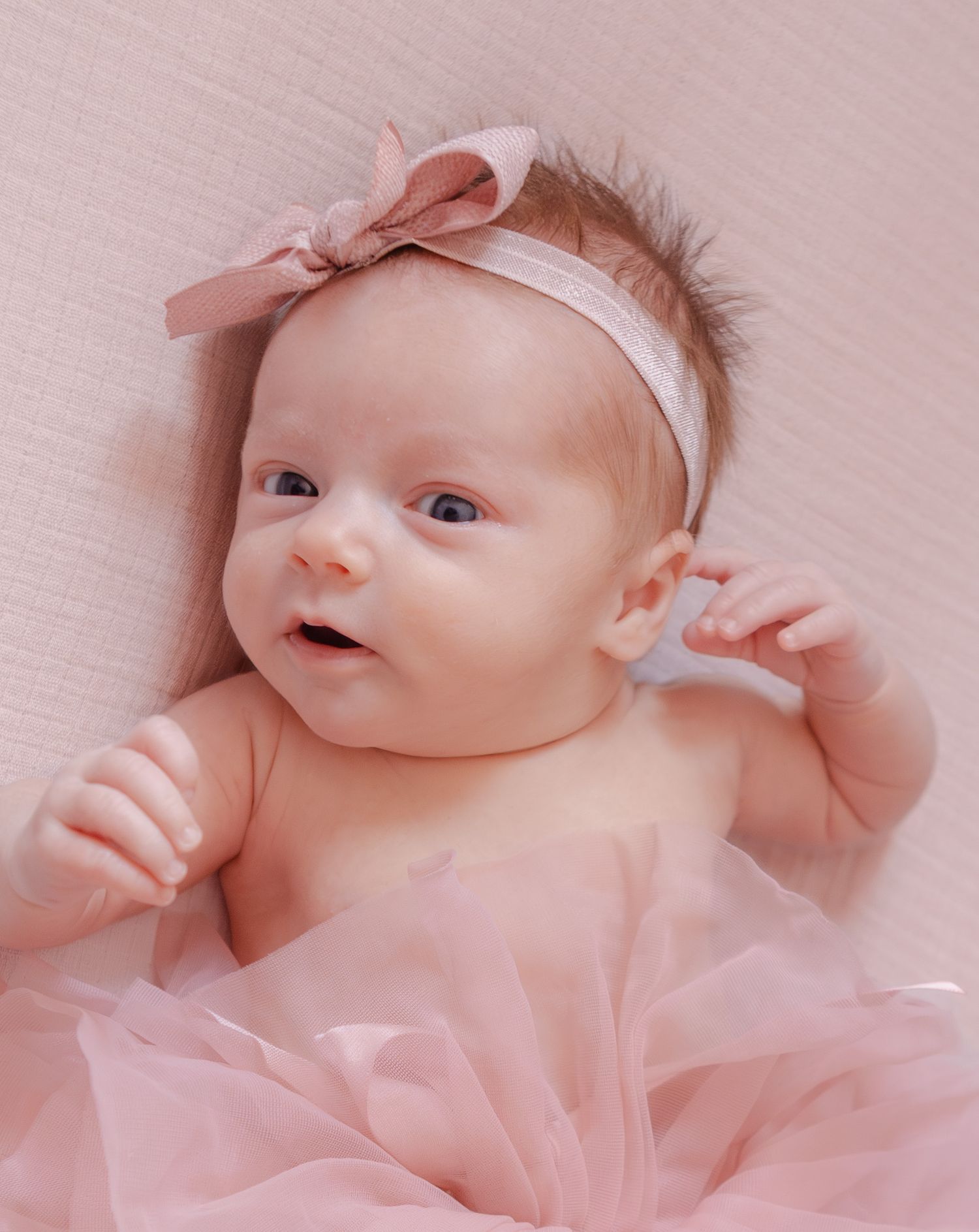 Adorable baby girl with pink bow headband and tutu, lying on pink background with bright eyes.