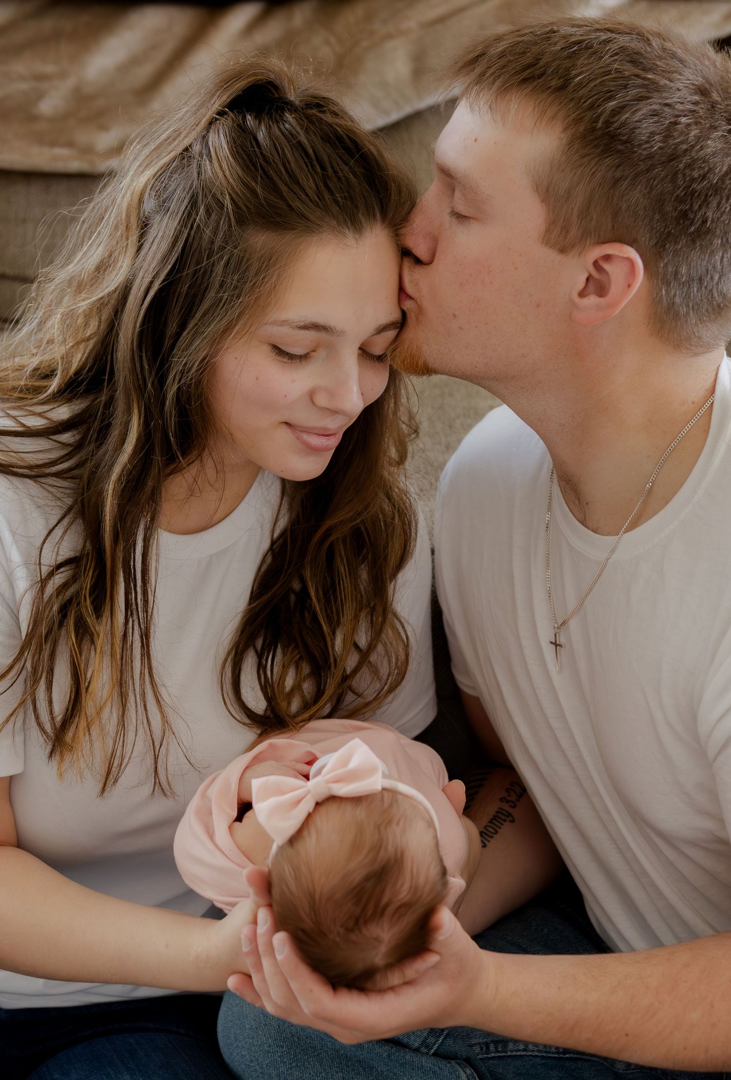 Young couple in white tees sharing a tender kiss while cradling their newborn baby girl with pink bow headband.