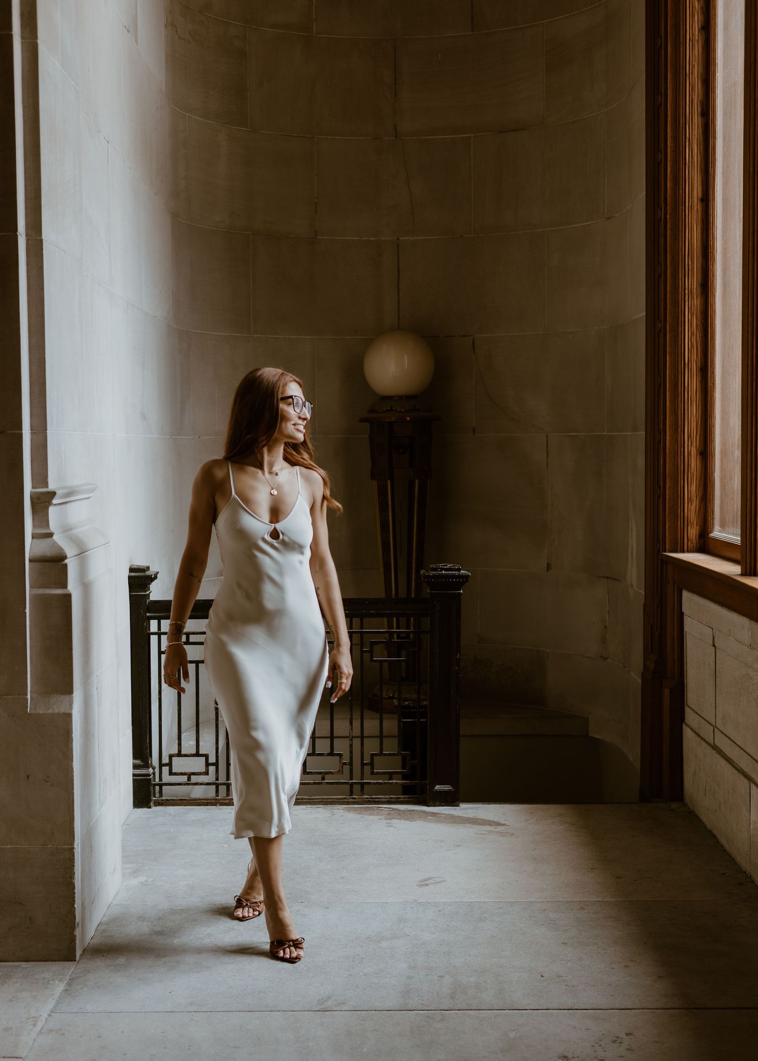 Elegant woman in a white satin slip dress walking through a grand architectural hallway.