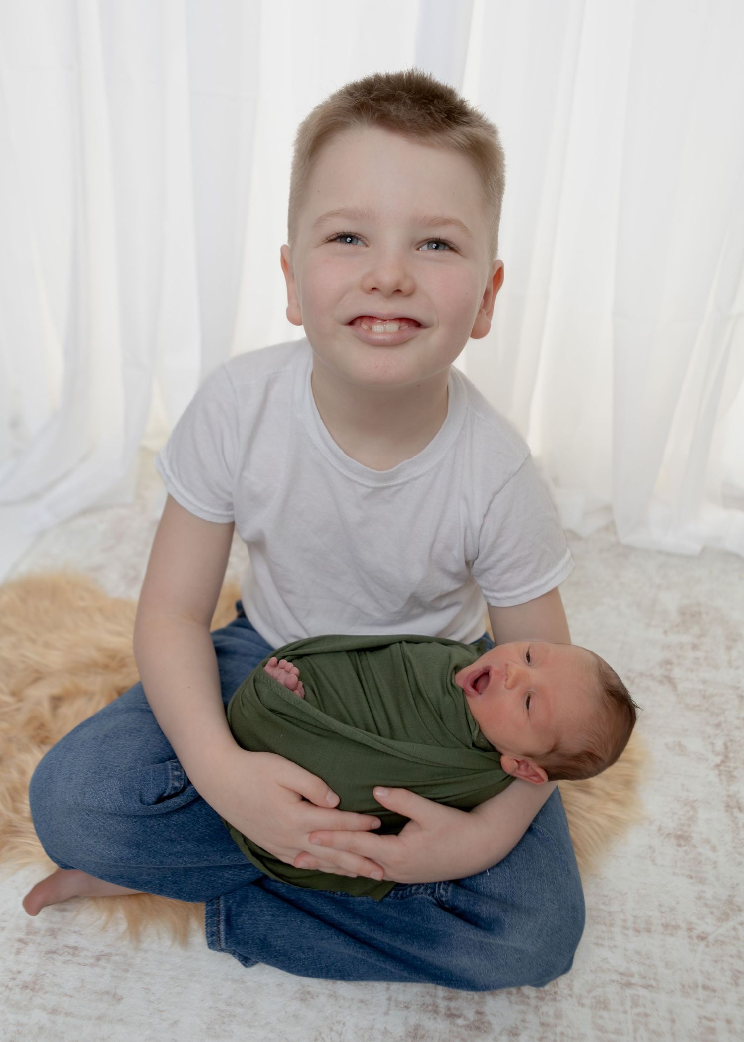 Smiling boy in white tee and jeans holds laughing newborn swaddled in green wrap.