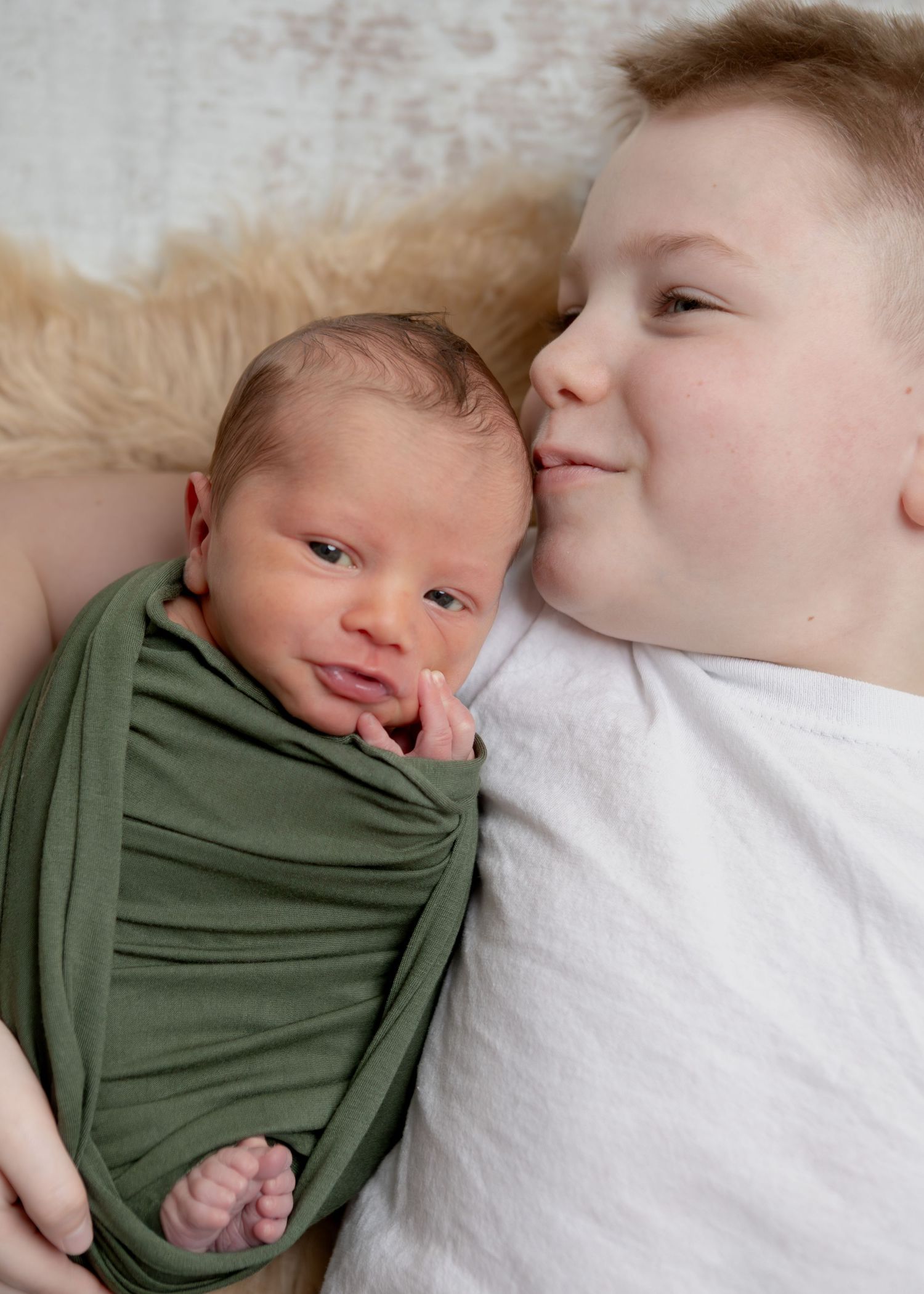 Older boy kisses a swaddled newborn baby wrapped in green fabric while lying on a white fur rug.