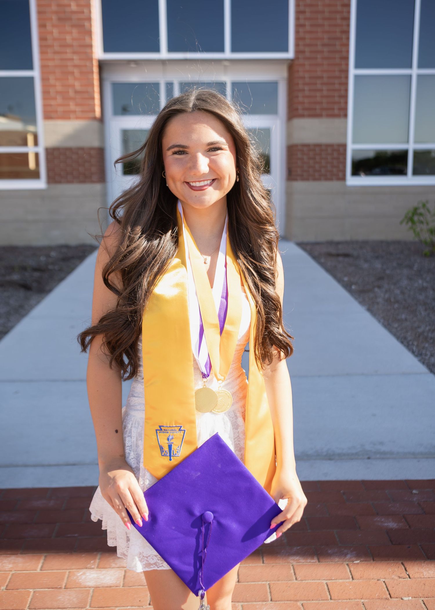 Smiling female graduate holds purple cap wearing gold honor stole and medal outside brick building.