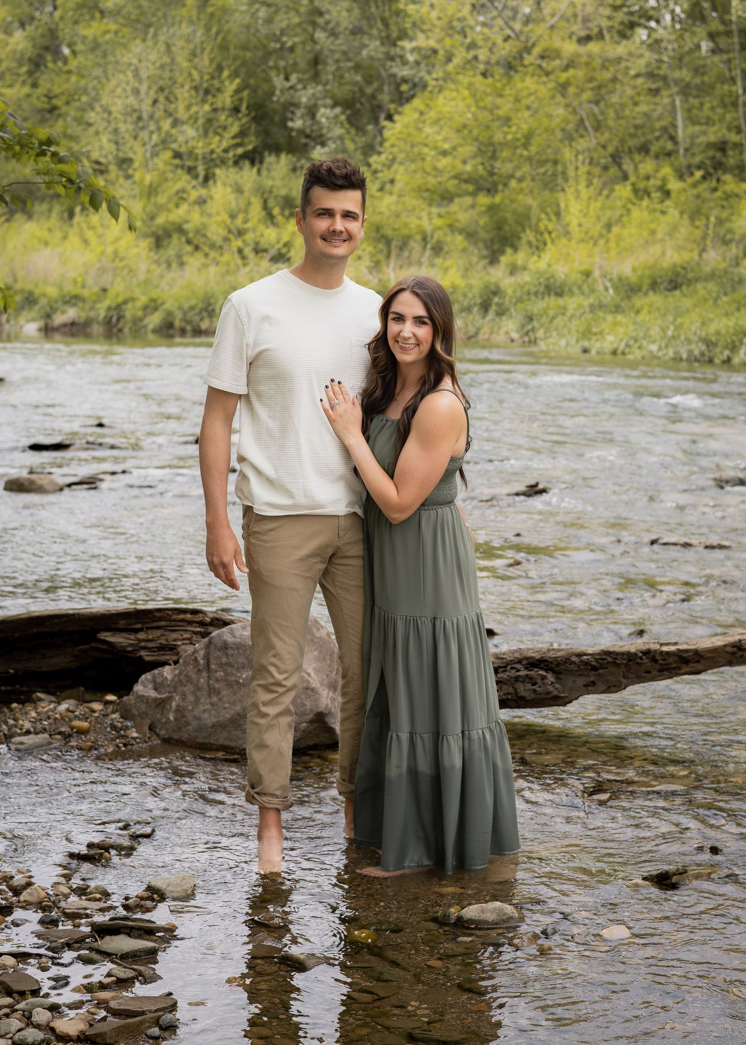 A couple stands barefoot in a rocky stream, smiling, surrounded by lush green trees.