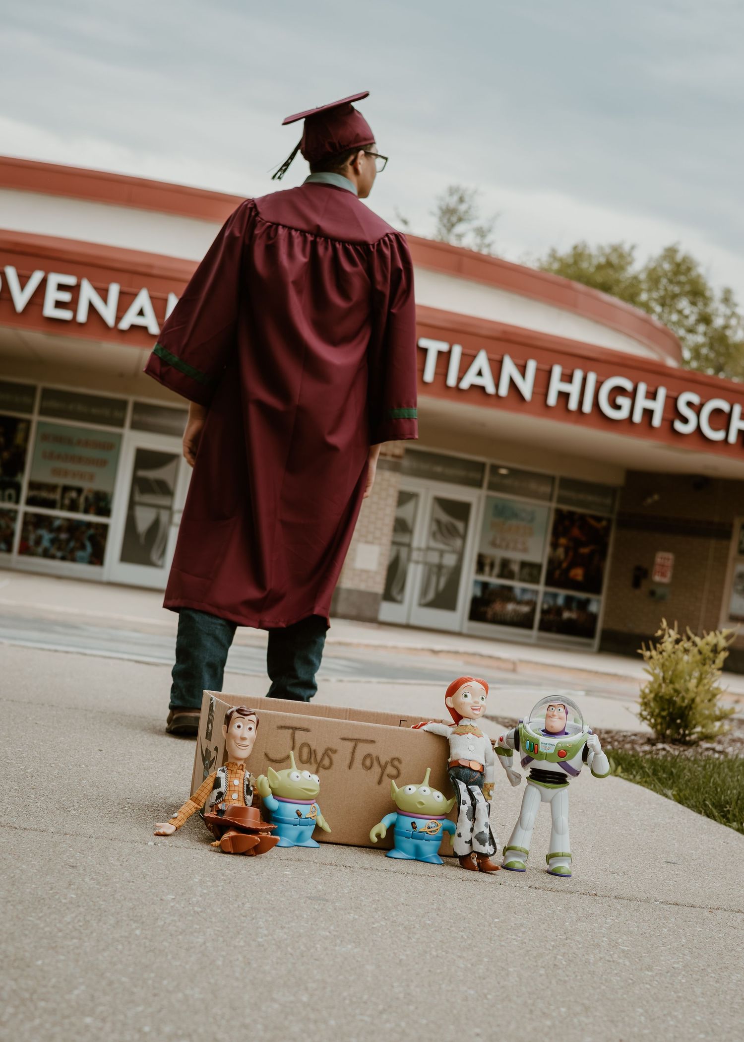 A graduate in maroon cap and gown stands before a high school with Toy Story figures displayed.