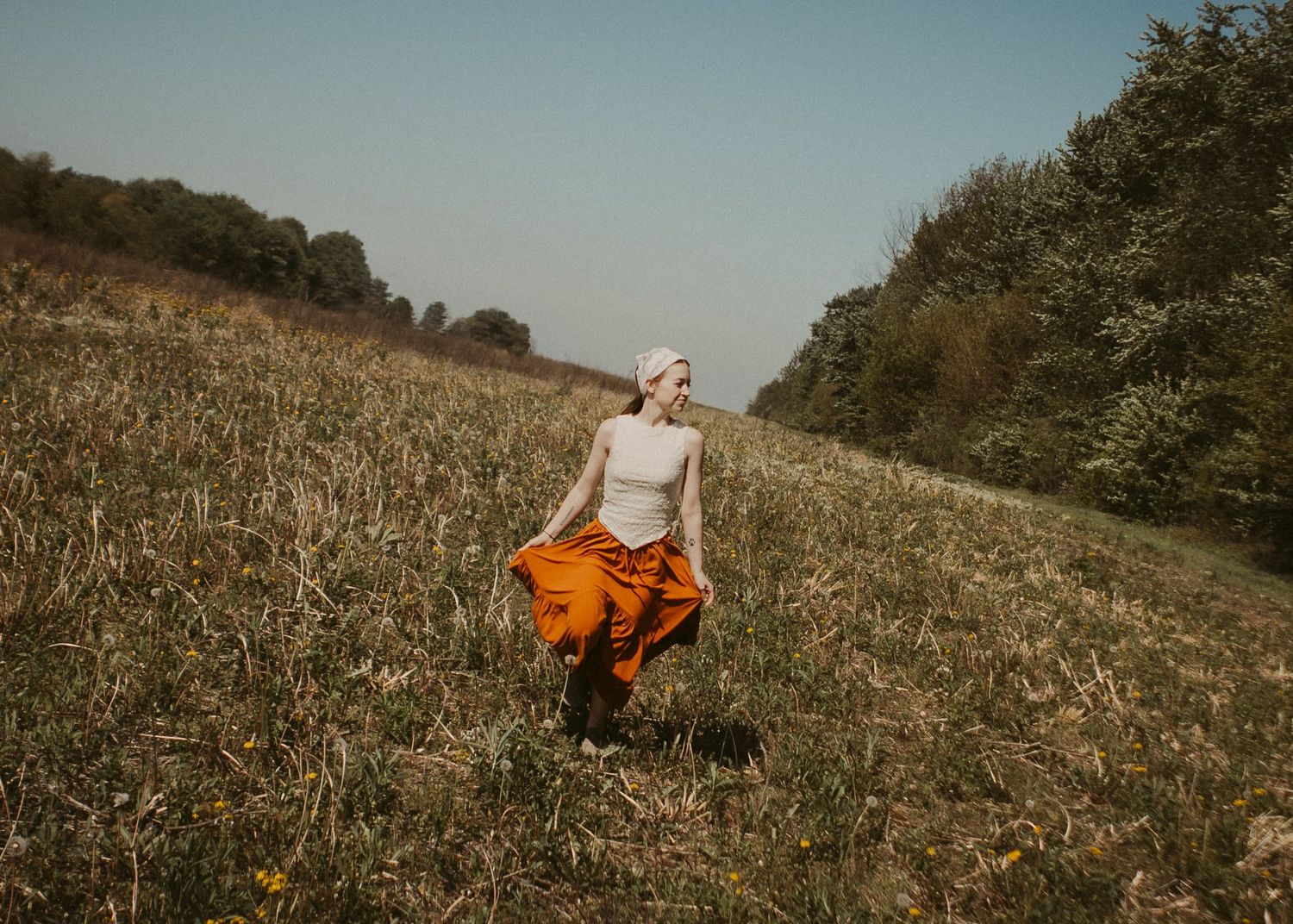 Woman in orange maxi skirt and white headscarf walking through a wildflower meadow.