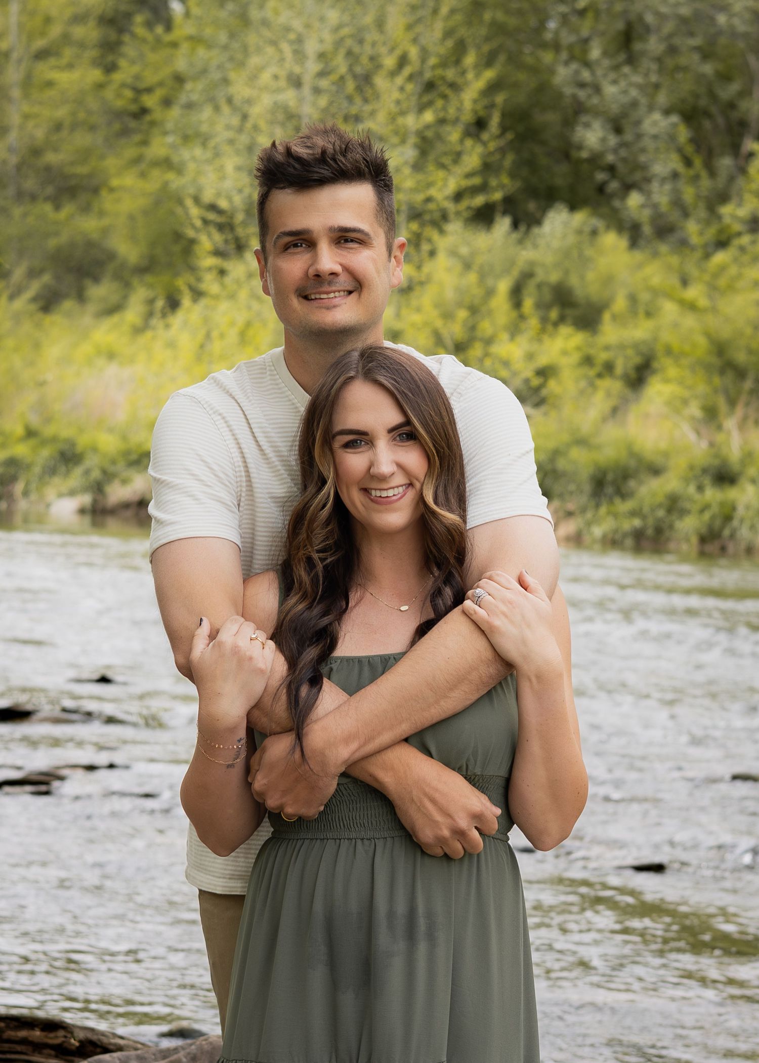 Happy couple embracing by a peaceful river surrounded by lush green trees.