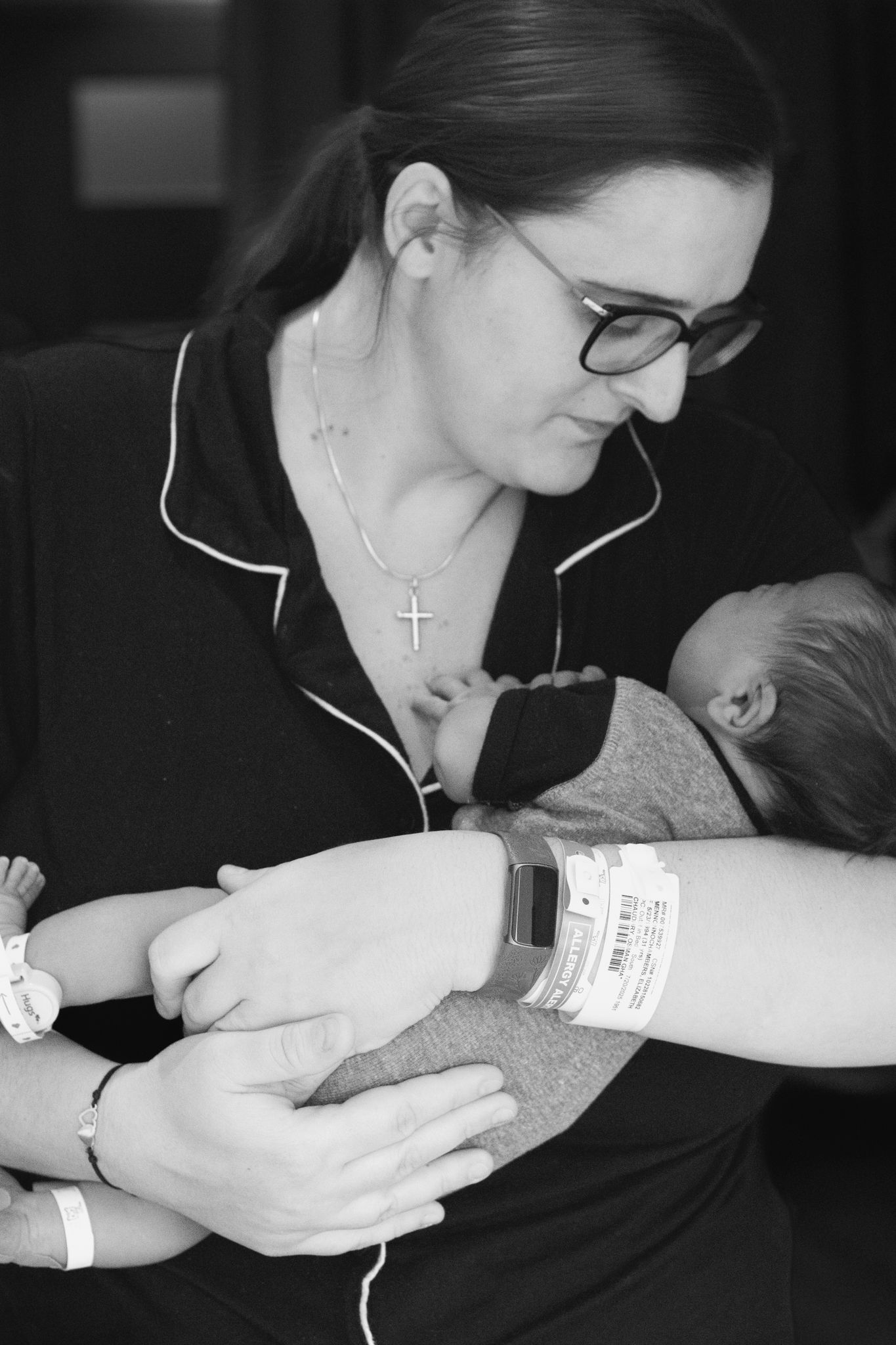 Black and white photo of a new mother wearing glasses and a cross necklace cradling her newborn in the hospital.
