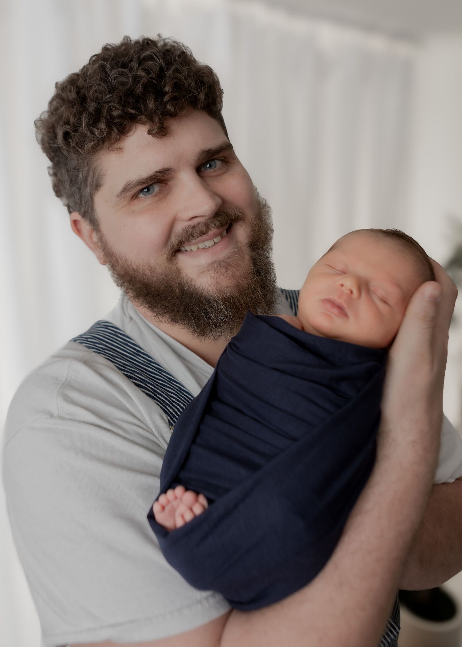 Smiling bearded father holds sleeping newborn swaddled in navy blue wrap against white backdrop.