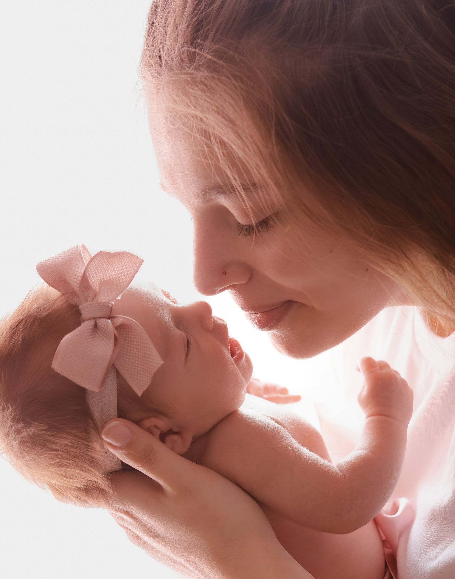 Mother tenderly holding newborn baby girl with pink bow headband close to her face in soft white light.