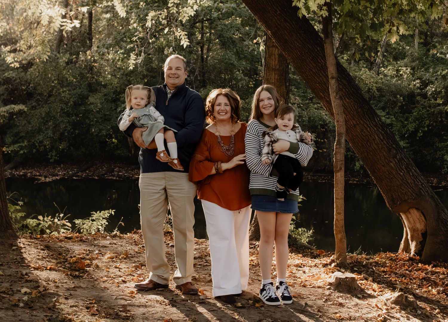 Grandparents and adult daughter holding two toddlers outdoors near a creek in fall foliage setting.