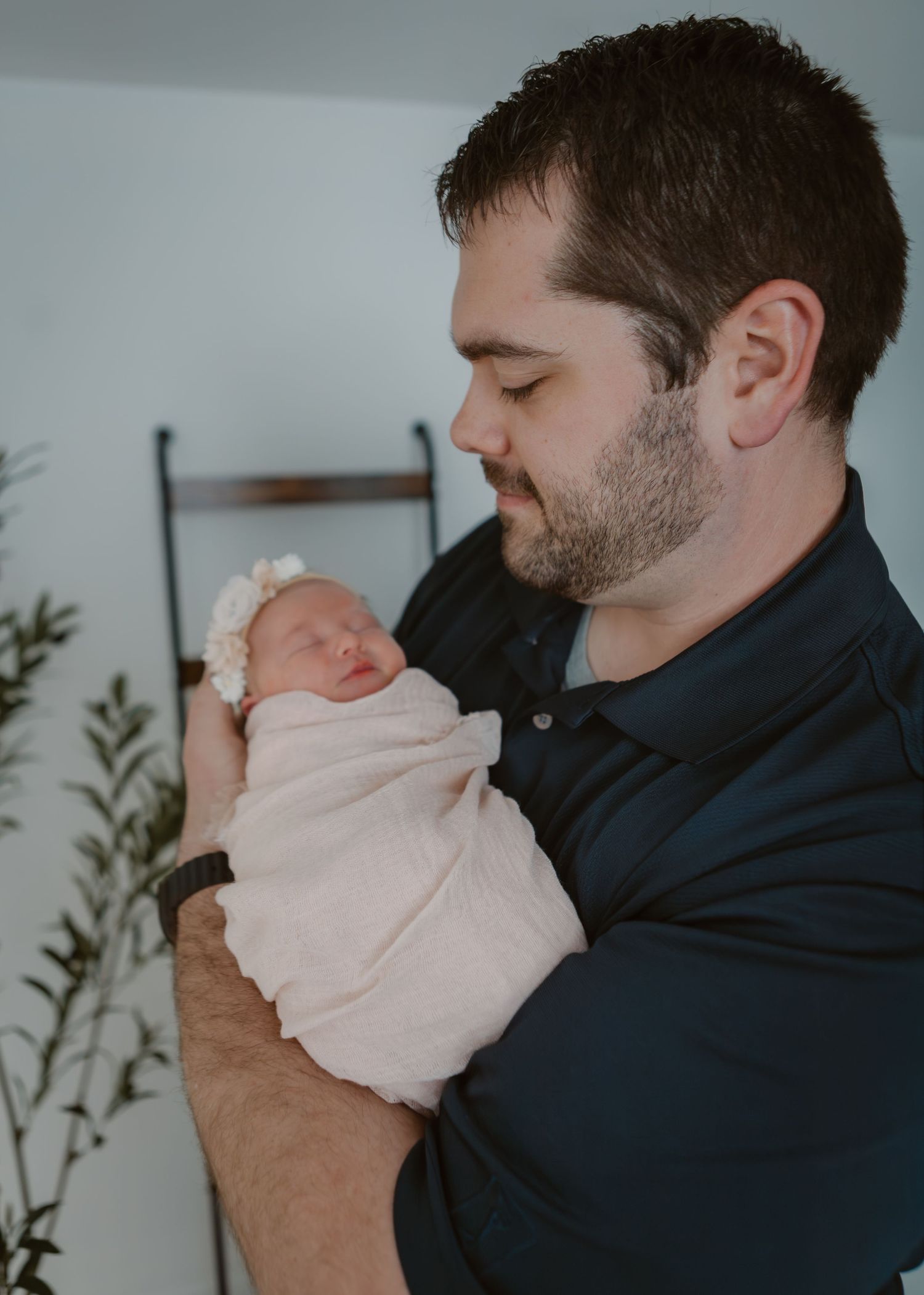 Father in navy shirt tenderly holding his sleeping newborn baby wrapped in white muslin.