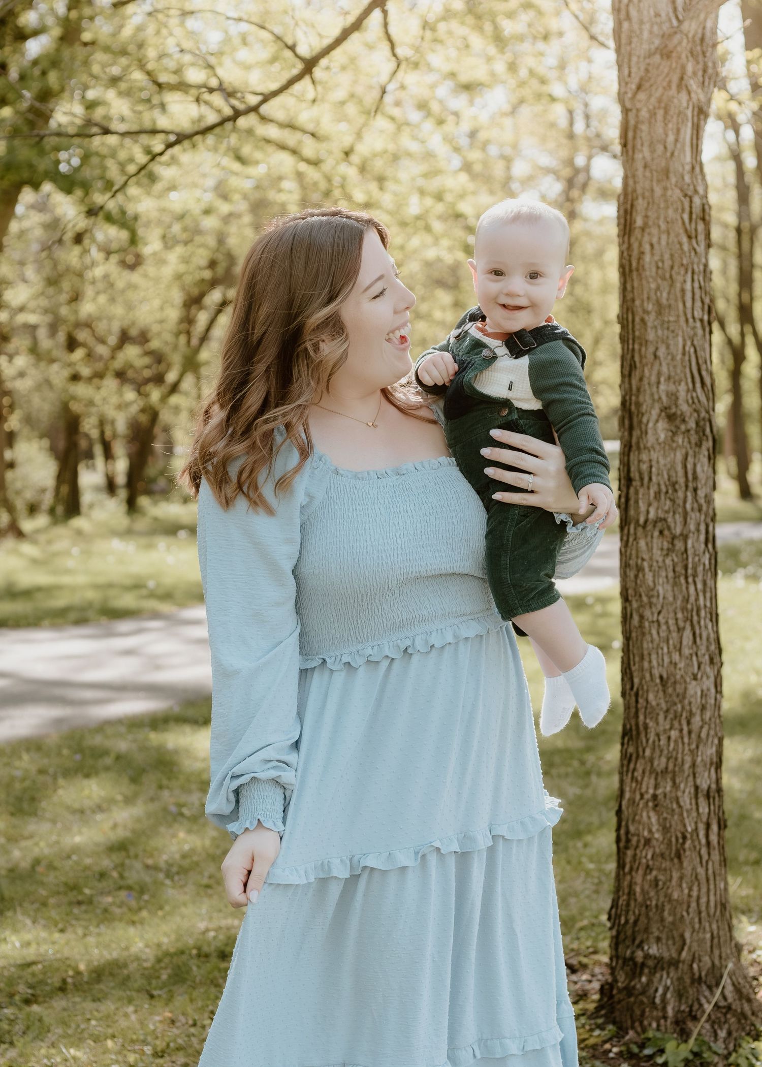 Laughing young mother in light blue smocked dress holding her baby boy outdoors among spring trees.