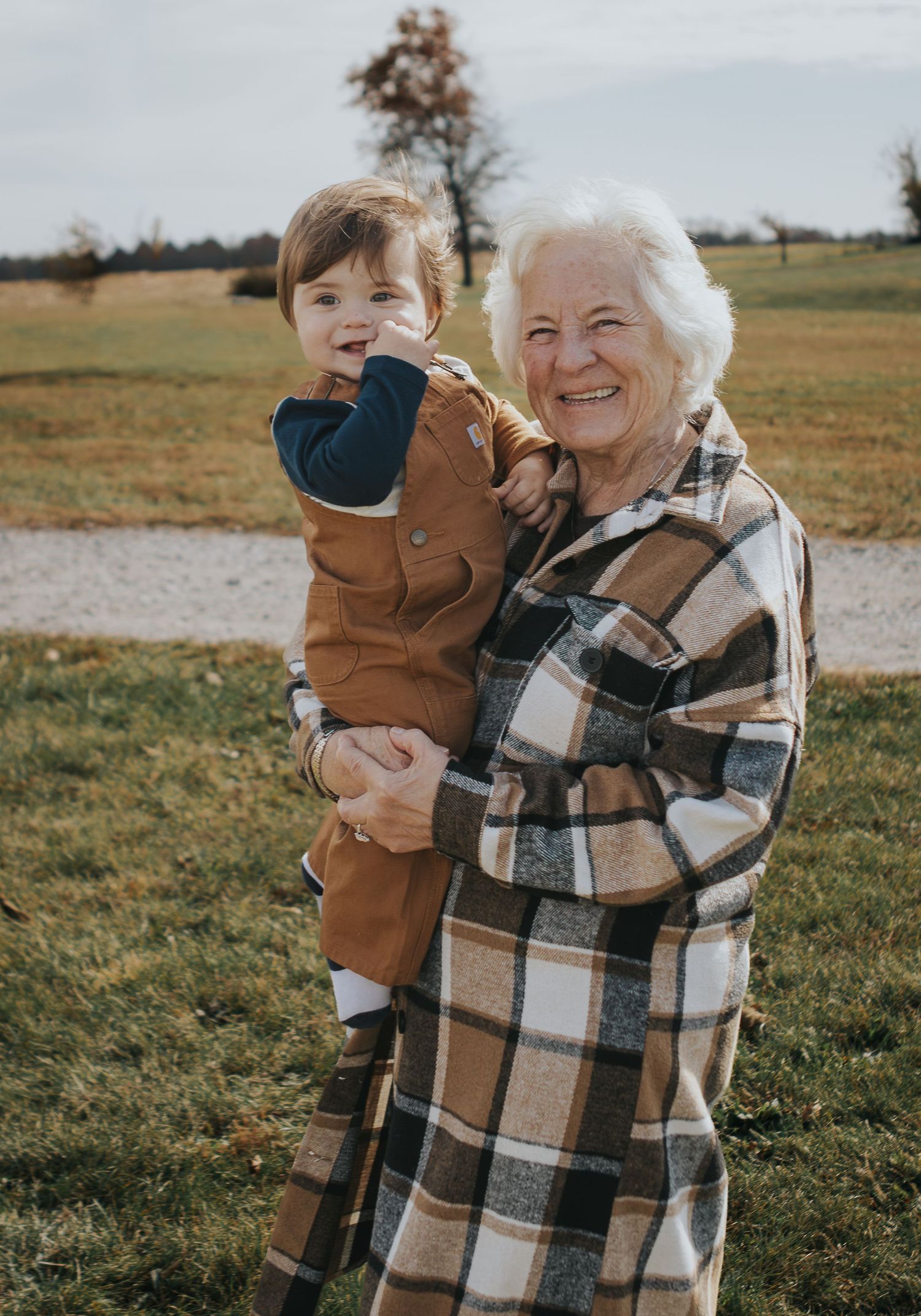 Elderly woman smiling warmly while holding a cheerful toddler in brown overalls outdoors in a grassy field.