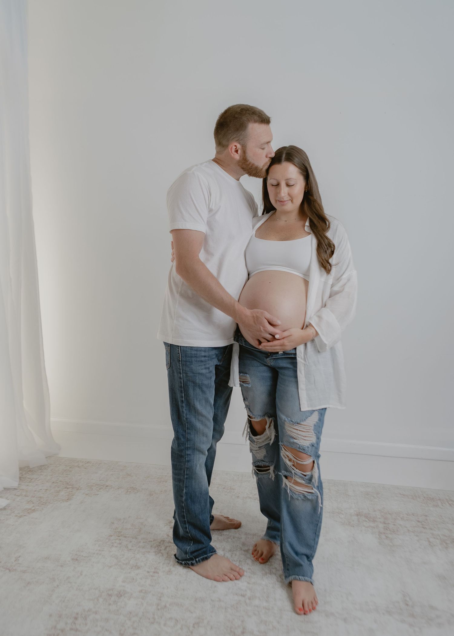Couple sharing a tender moment, man kissing pregnant woman's head in a bright studio.