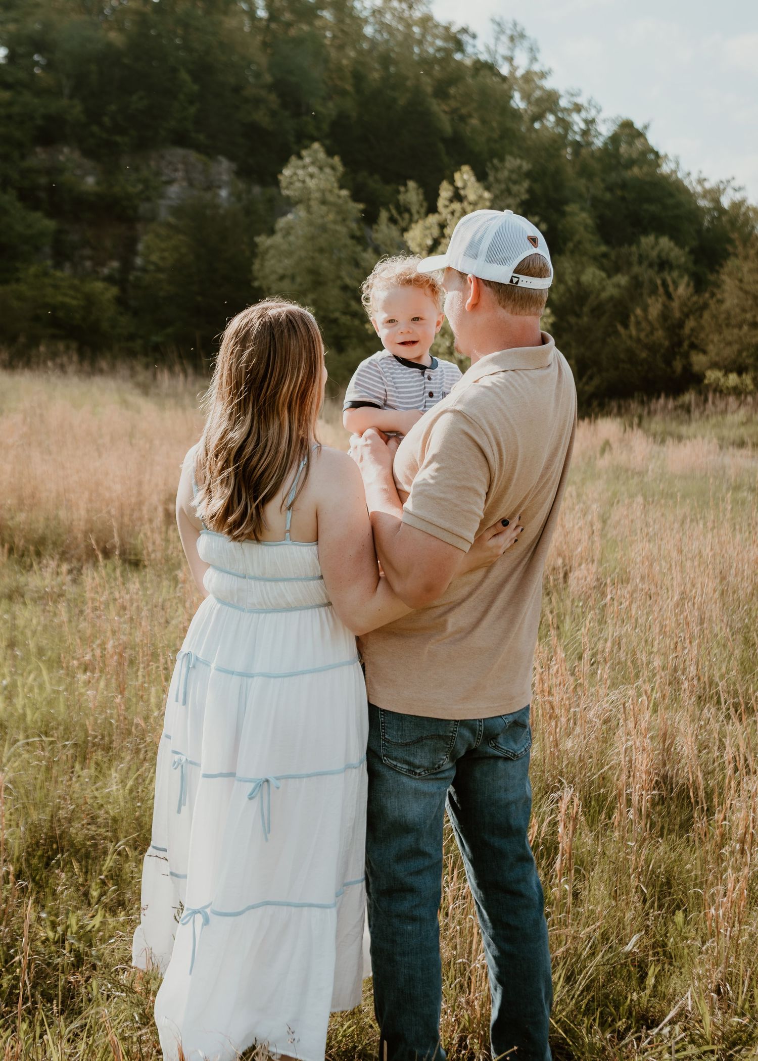 Young couple holding toddler boy in golden meadow during outdoor family photo session.