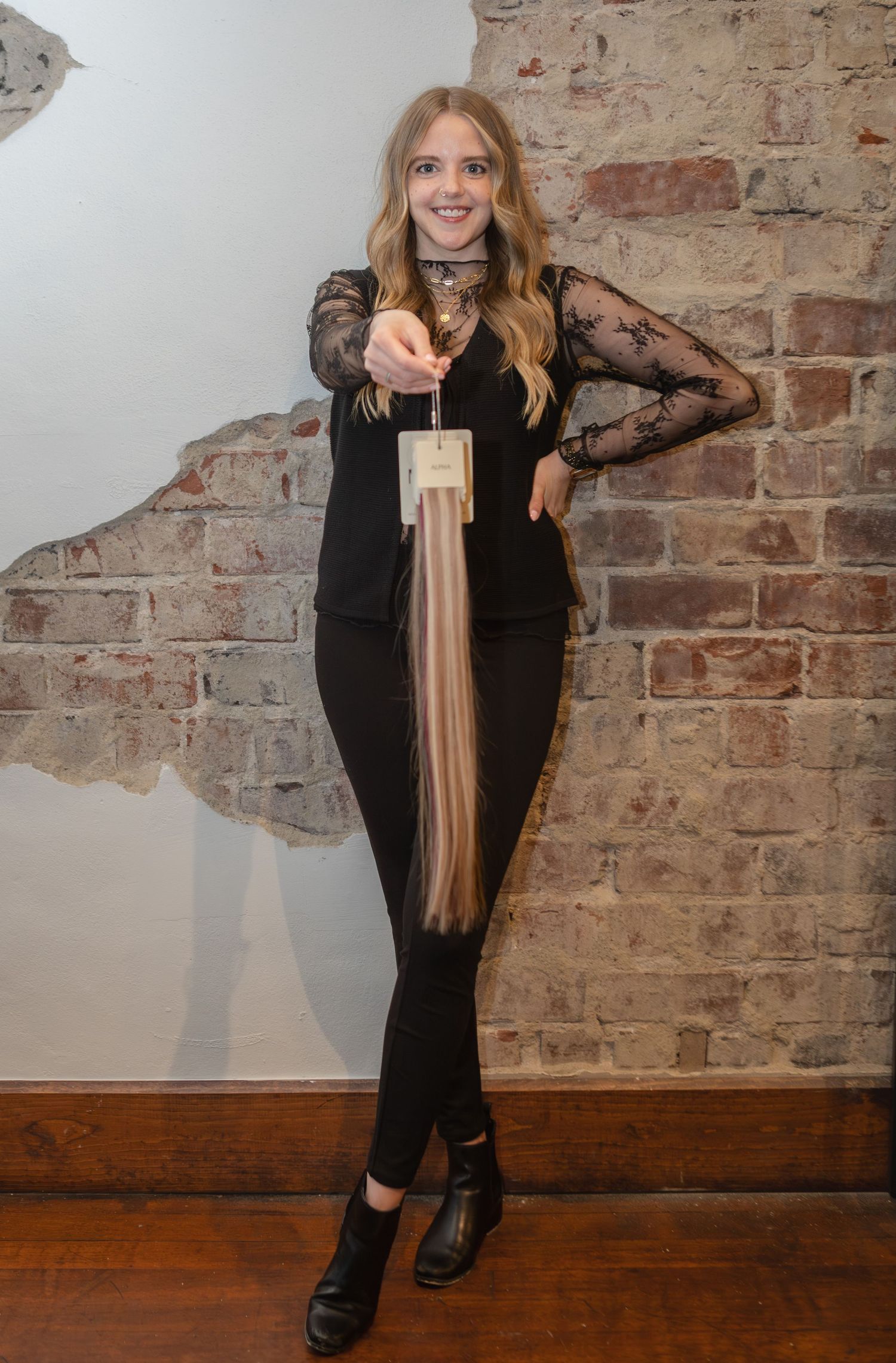 Young blonde woman in black outfit holds hair extensions toward camera in front of exposed brick wall.