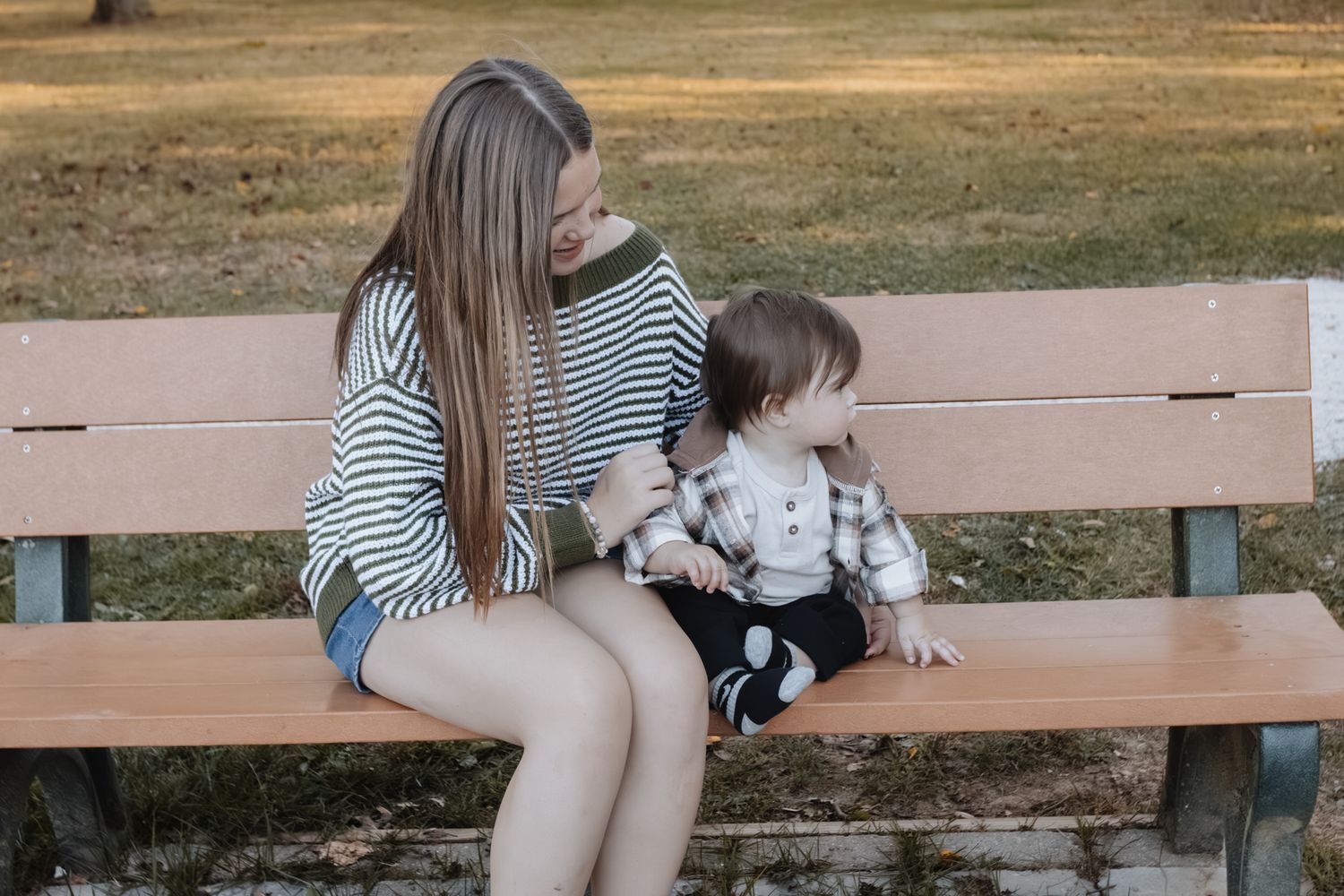 Young mother sitting on park bench with toddler in striped shirts on sunny day outdoors.