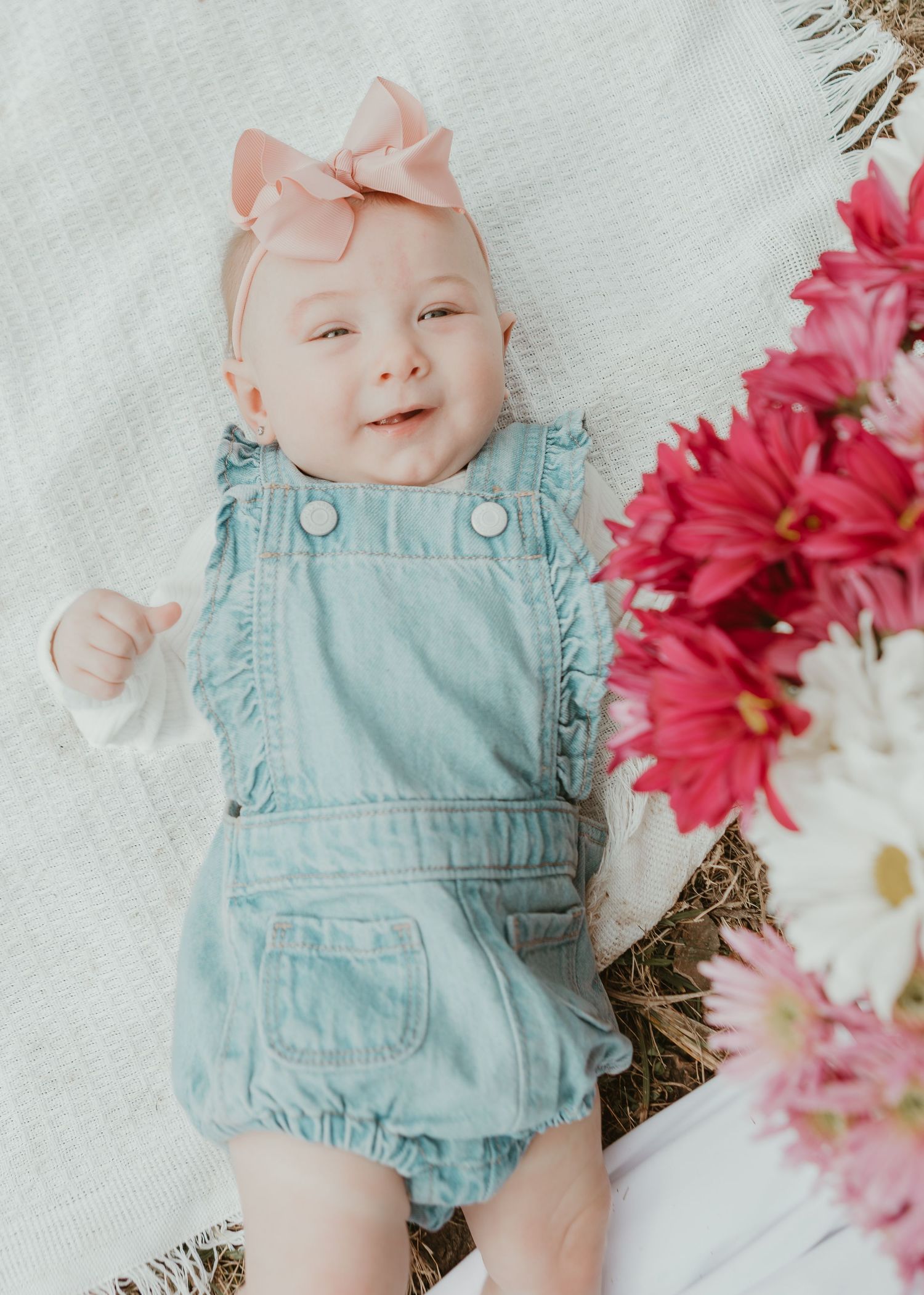 Adorable baby girl in denim romper and pink bow lying beside vibrant spring flowers.