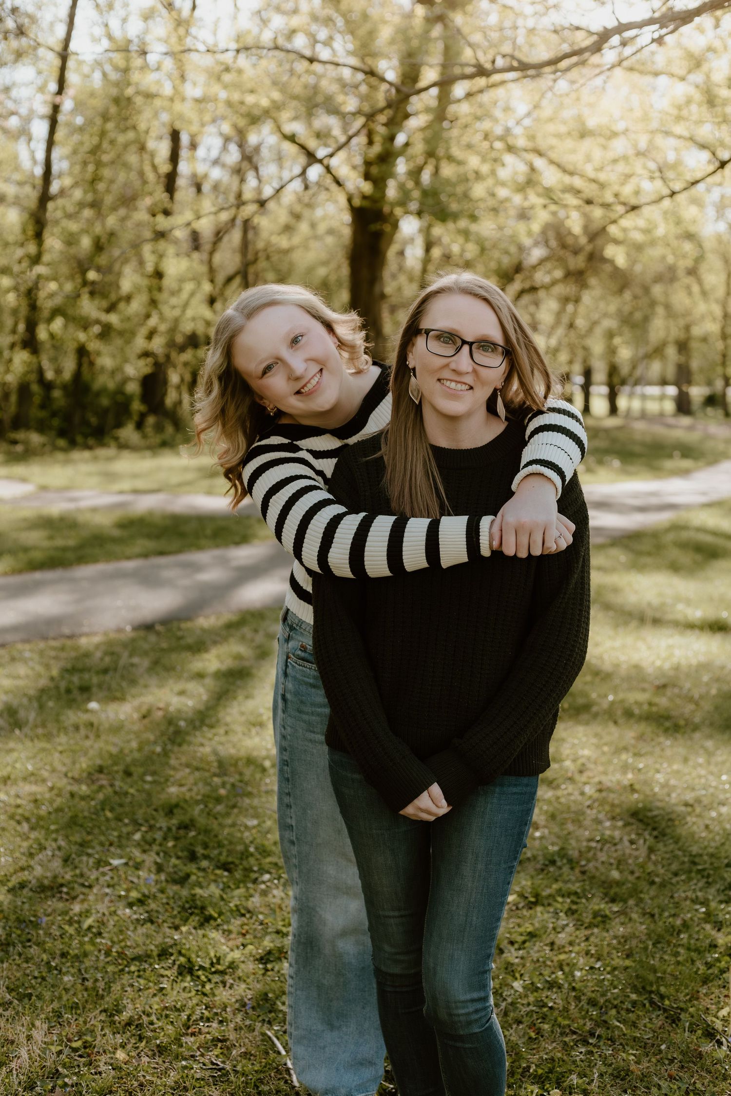 Two teenage girls hugging and smiling together outdoors in a tree-lined park.