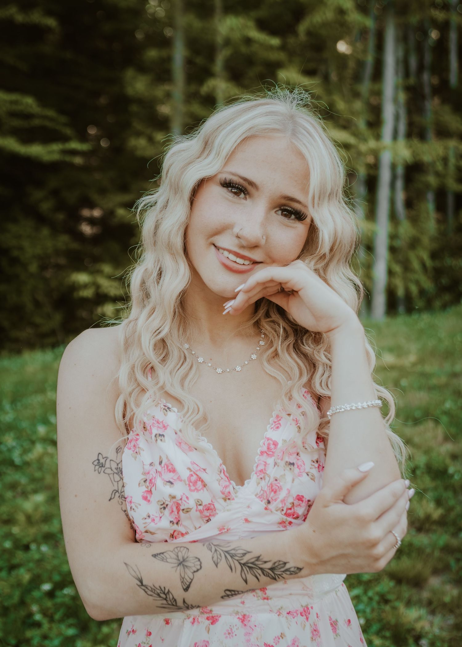 Smiling blonde woman in pink floral dress with tattoo posing in front of green forest.