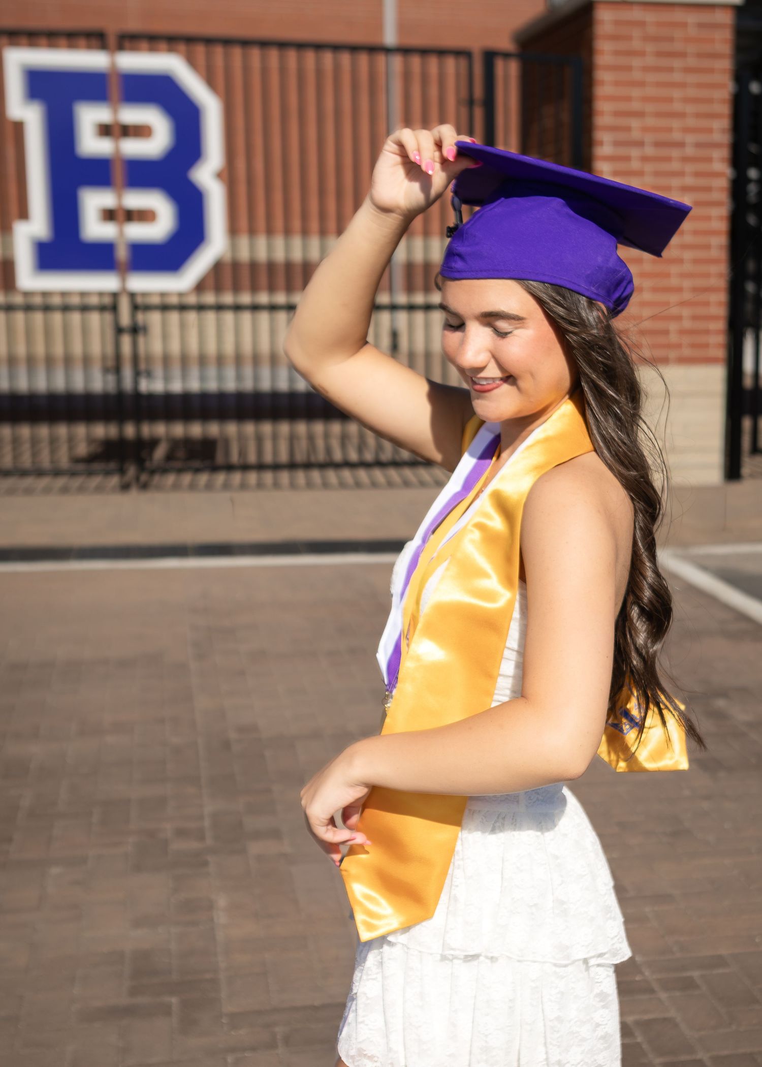 Young woman in purple graduation cap and gold honor stole poses outdoors near brick building with letter B.