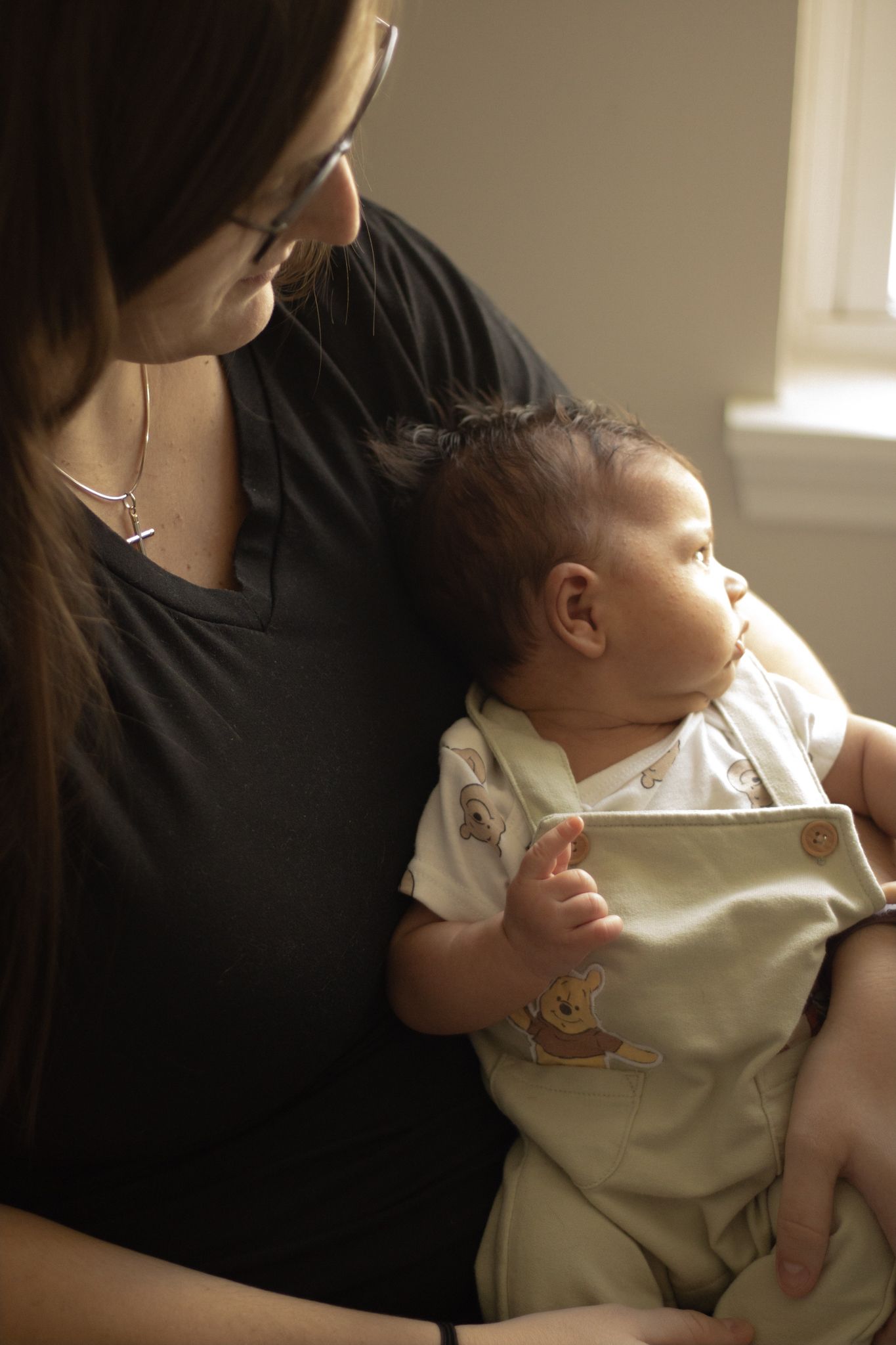 Mother with glasses in black shirt gently holding her newborn baby dressed in Winnie the Pooh overalls.