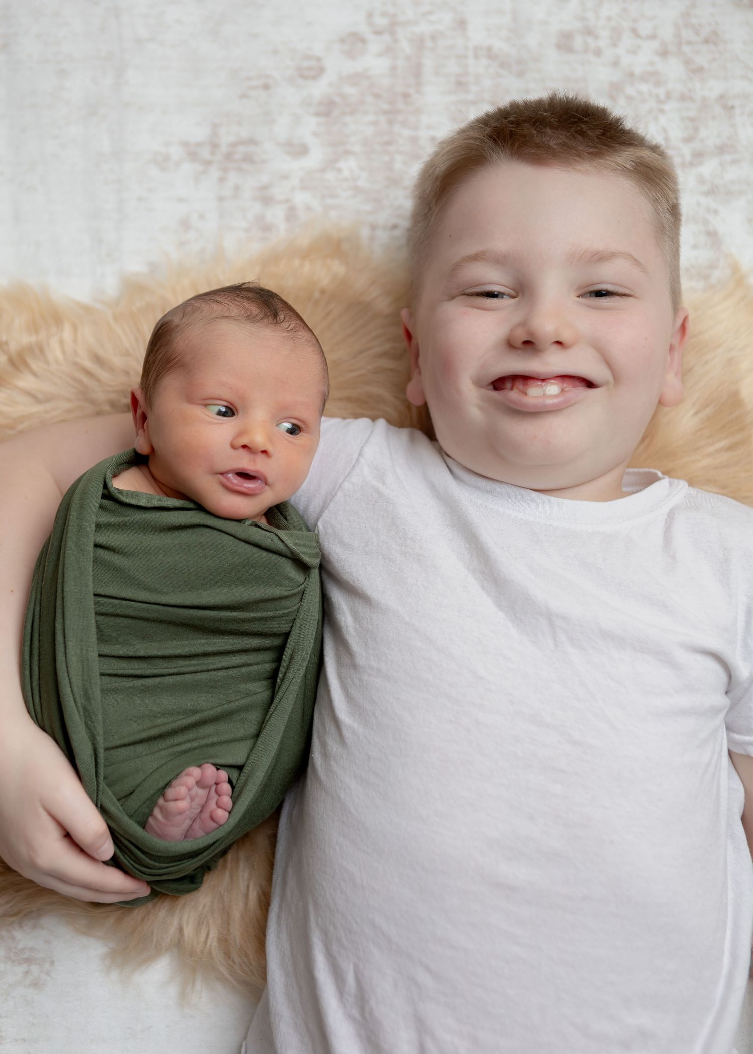 Smiling boy lies beside newborn baby swaddled in green wrap on fluffy fur backdrop.