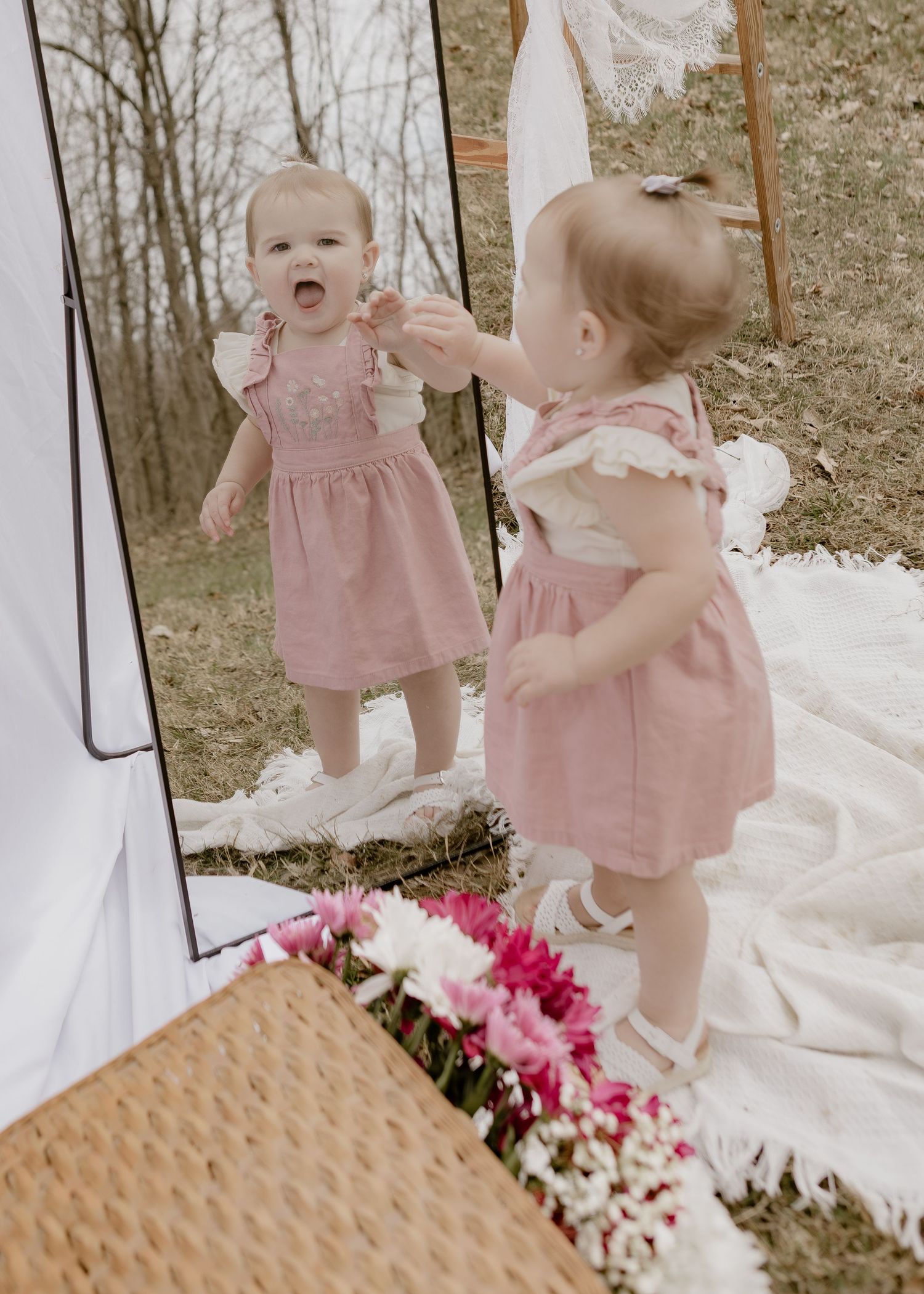 Curious baby girl in pink dress discovering her reflection in a tall mirror outdoors.