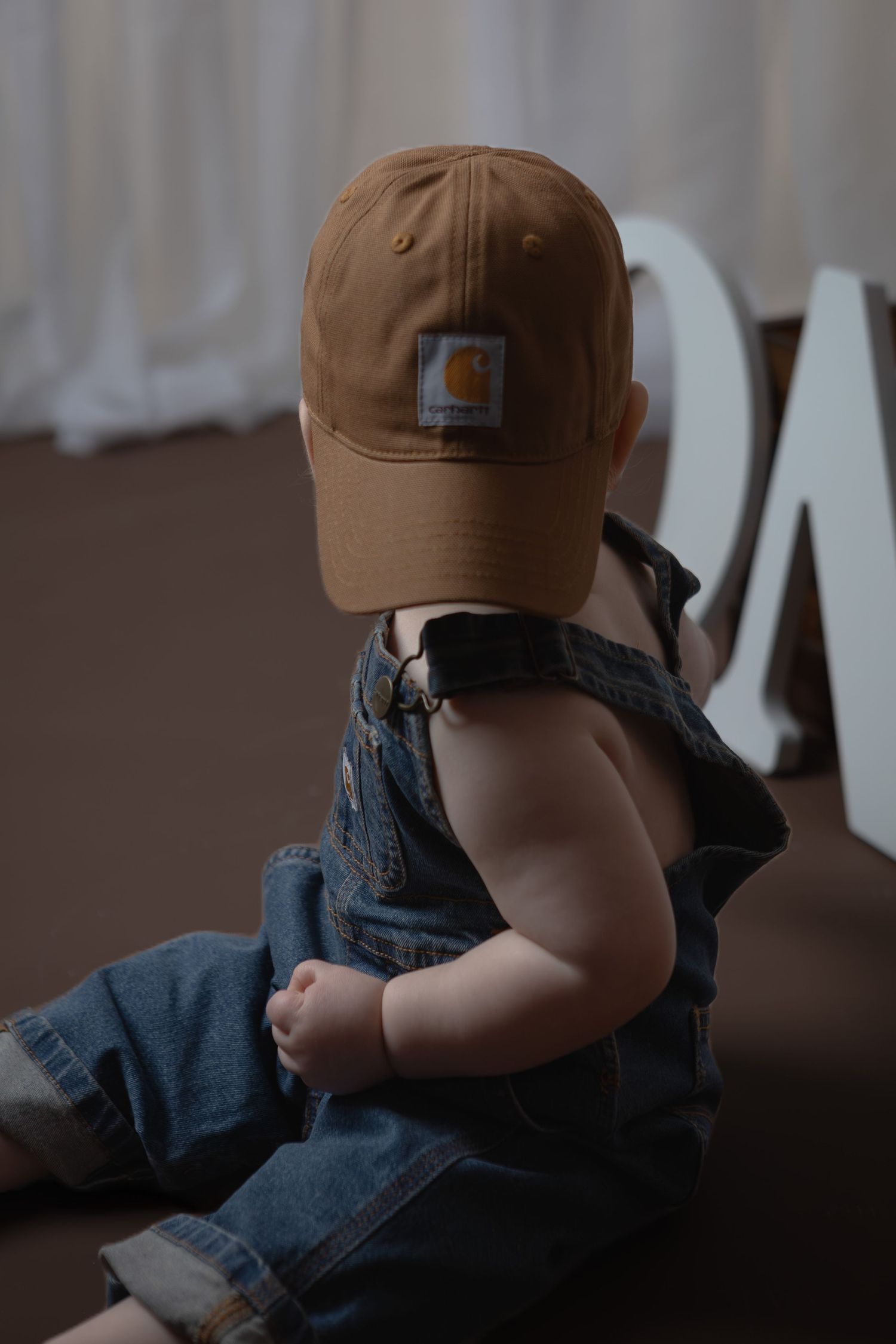 Adorable baby wearing Carhartt cap and denim overalls sitting in front of white letter decor.