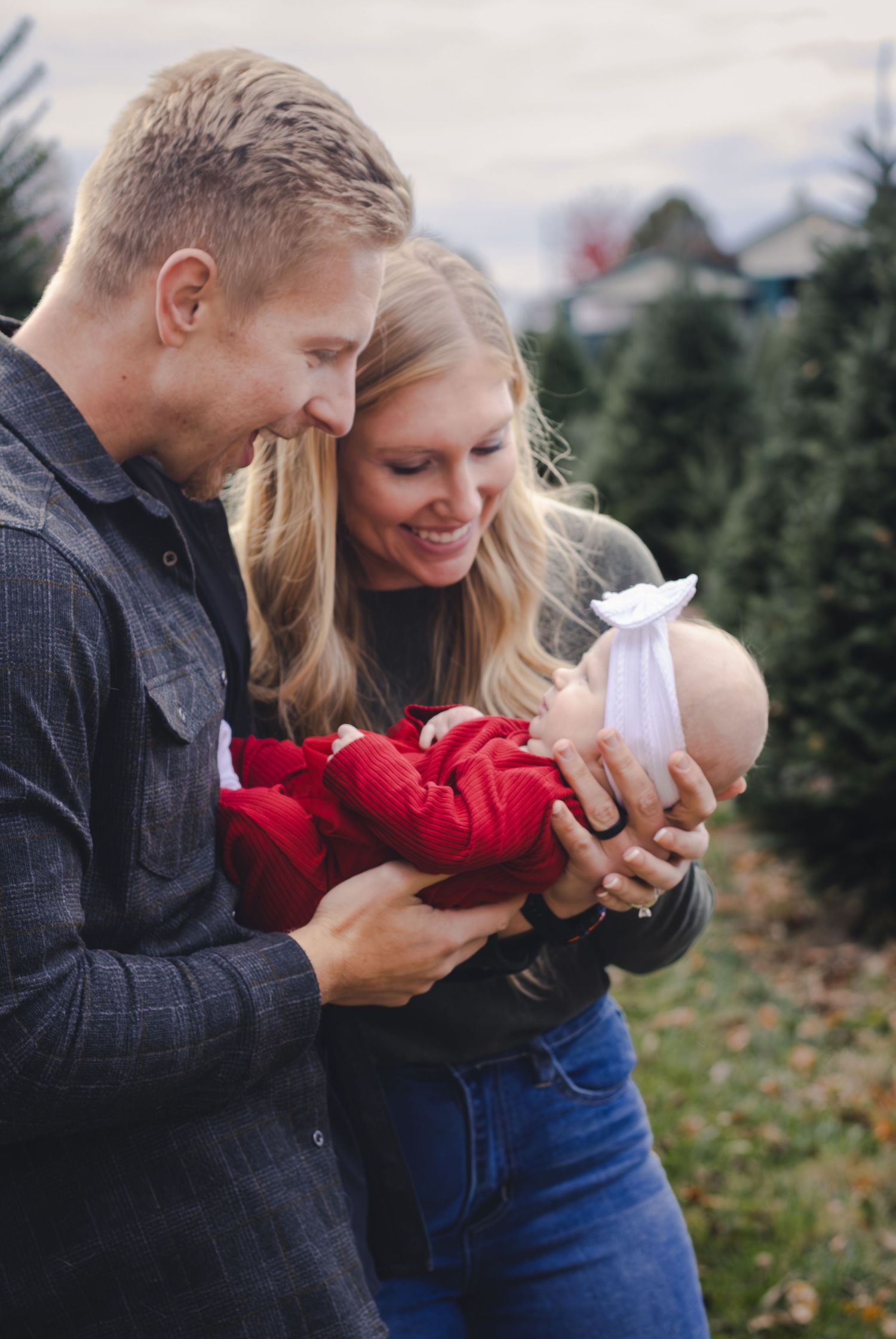 A couple smiles lovingly at their newborn baby dressed in red at an outdoor Christmas tree farm.
