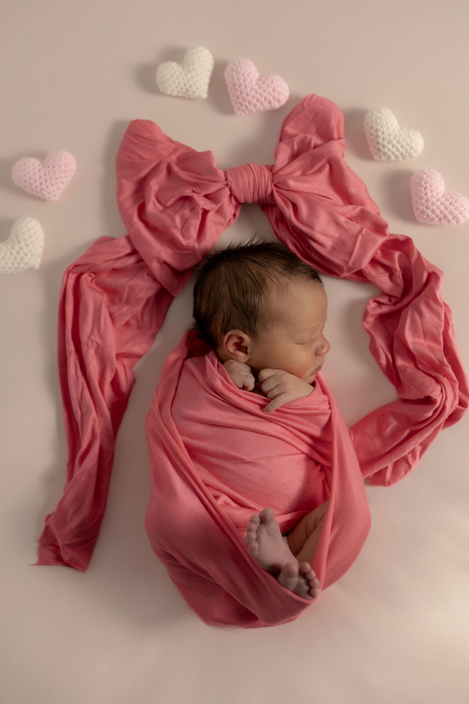 Sleeping newborn baby swaddled in pink fabric with a bow, surrounded by crocheted hearts on a white background.
