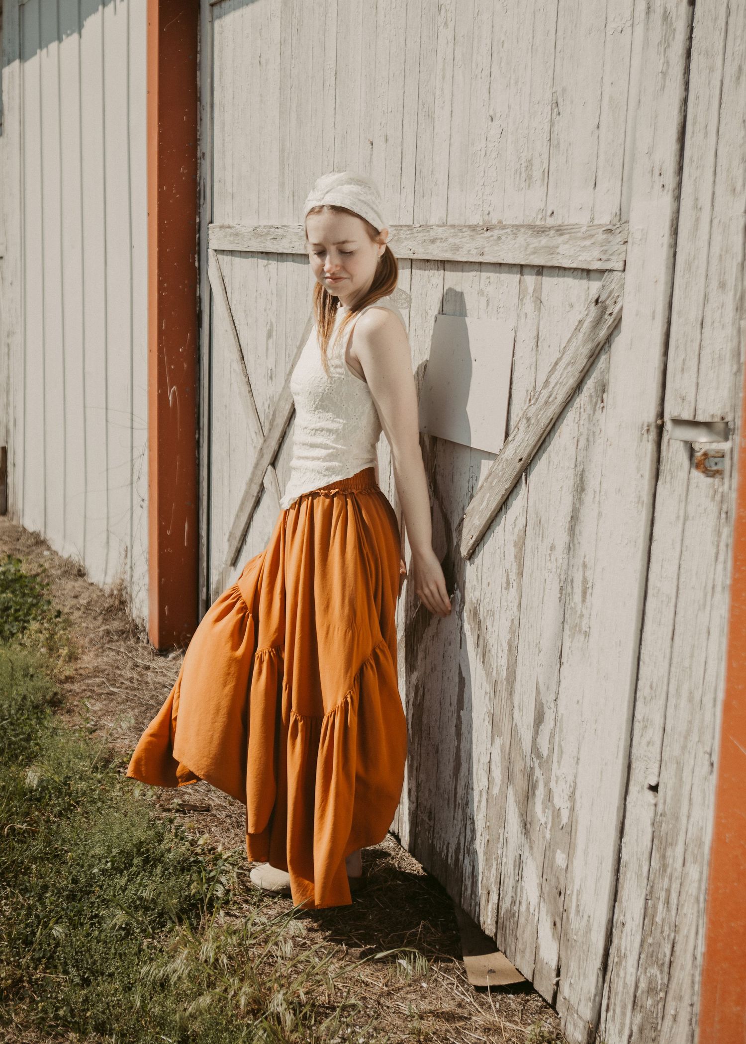 Woman in orange tiered maxi skirt and white tank top leaning against rustic barn door outdoors.