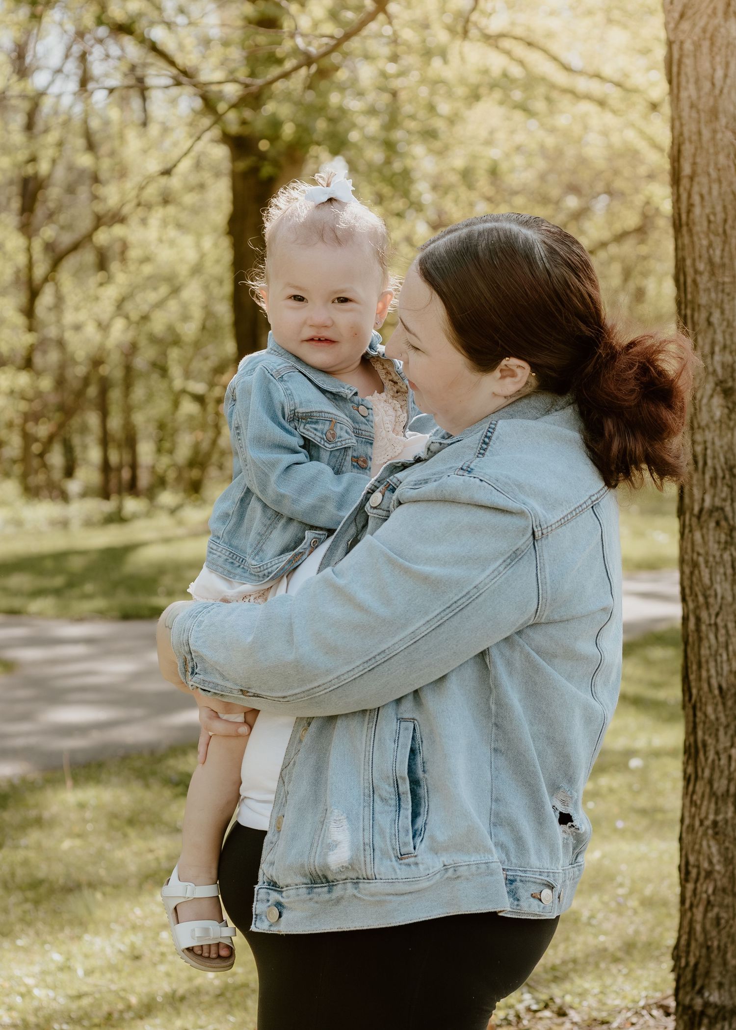 Mother in denim jacket holding toddler daughter outdoors in a sunny wooded park.
