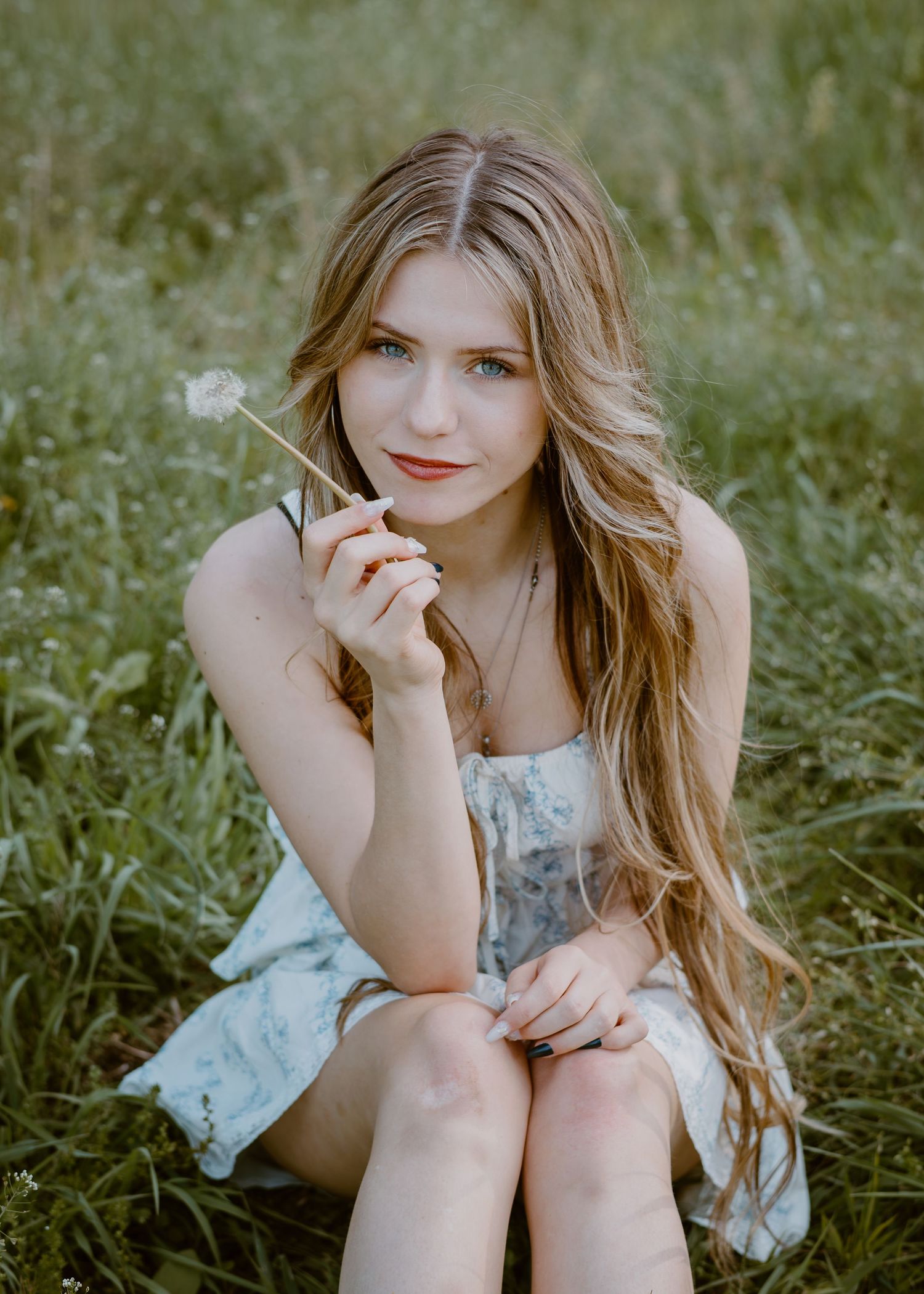 Young woman with long wavy hair sitting in grass field holding a dandelion, wearing a light floral dress.