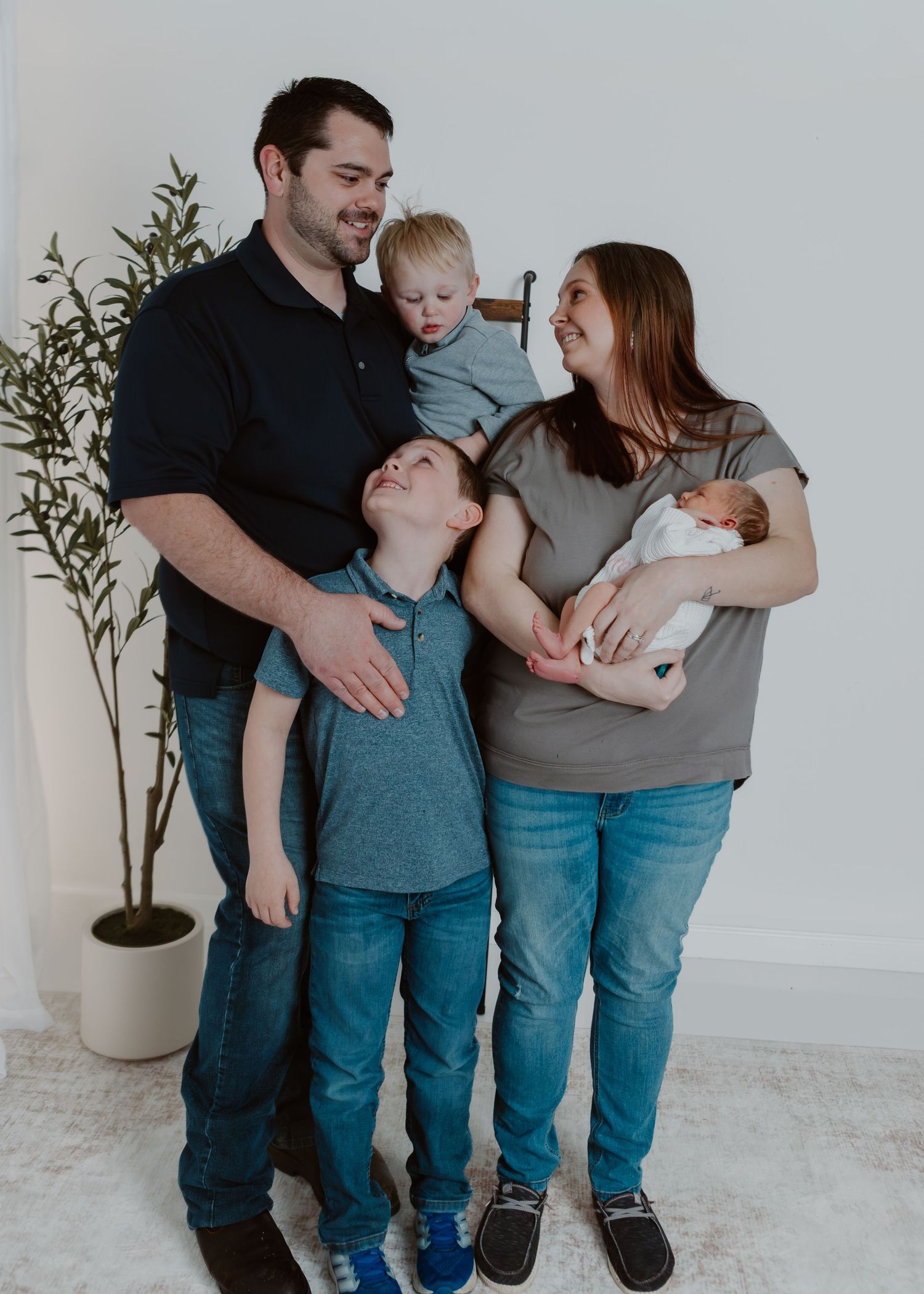 Happy family of five posing indoors with newborn baby, toddler, and young boy smiling together.