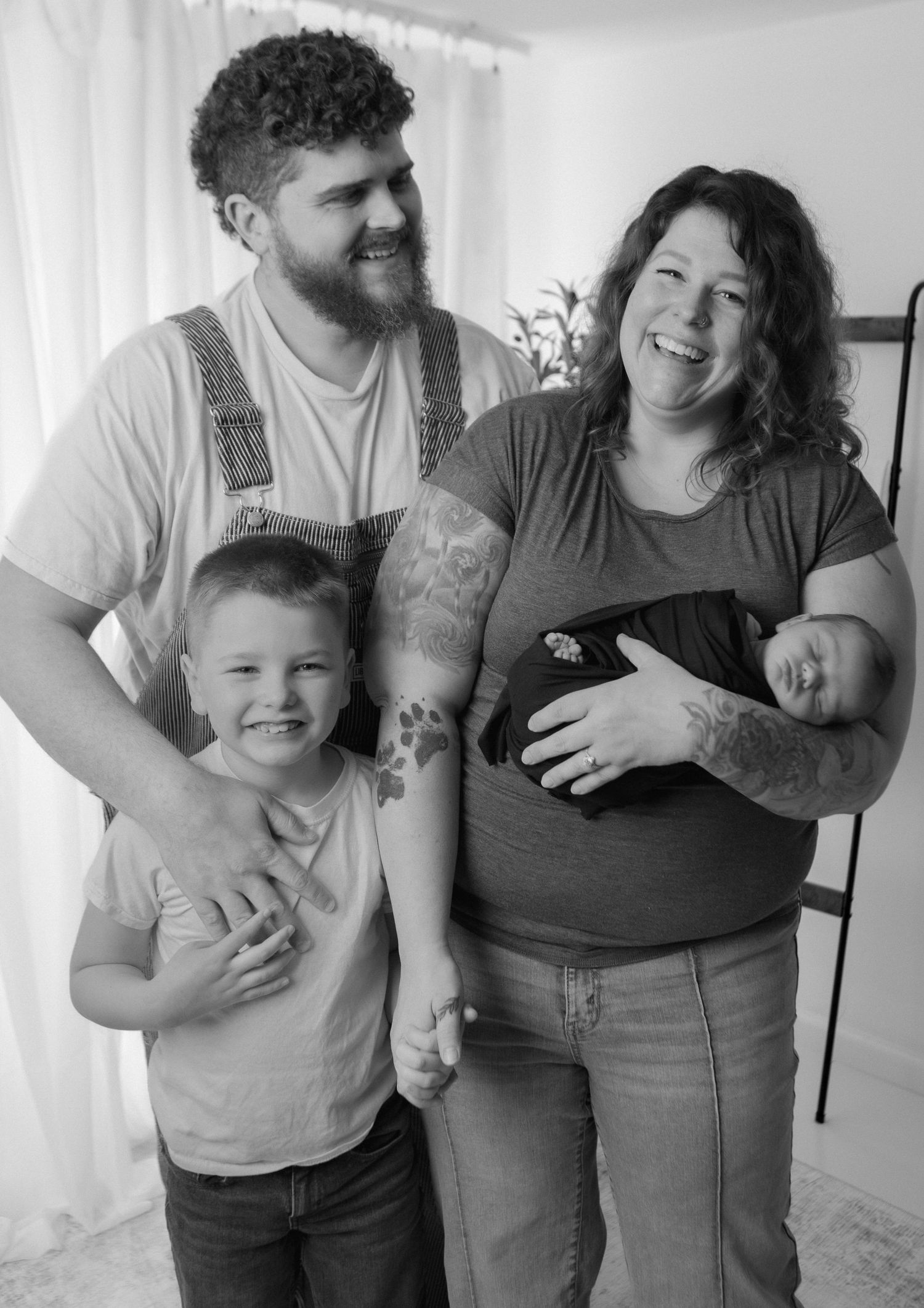 Black and white photo of a smiling family with tattooed parents, young child, and newborn baby.