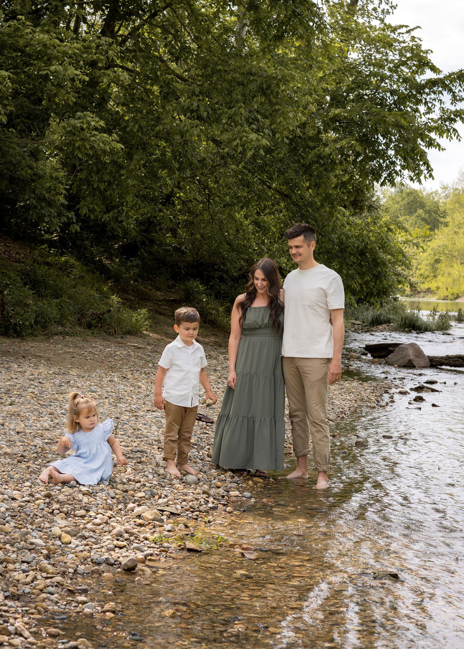 Family of four posing together on a rocky riverbank with tall green trees in the background.