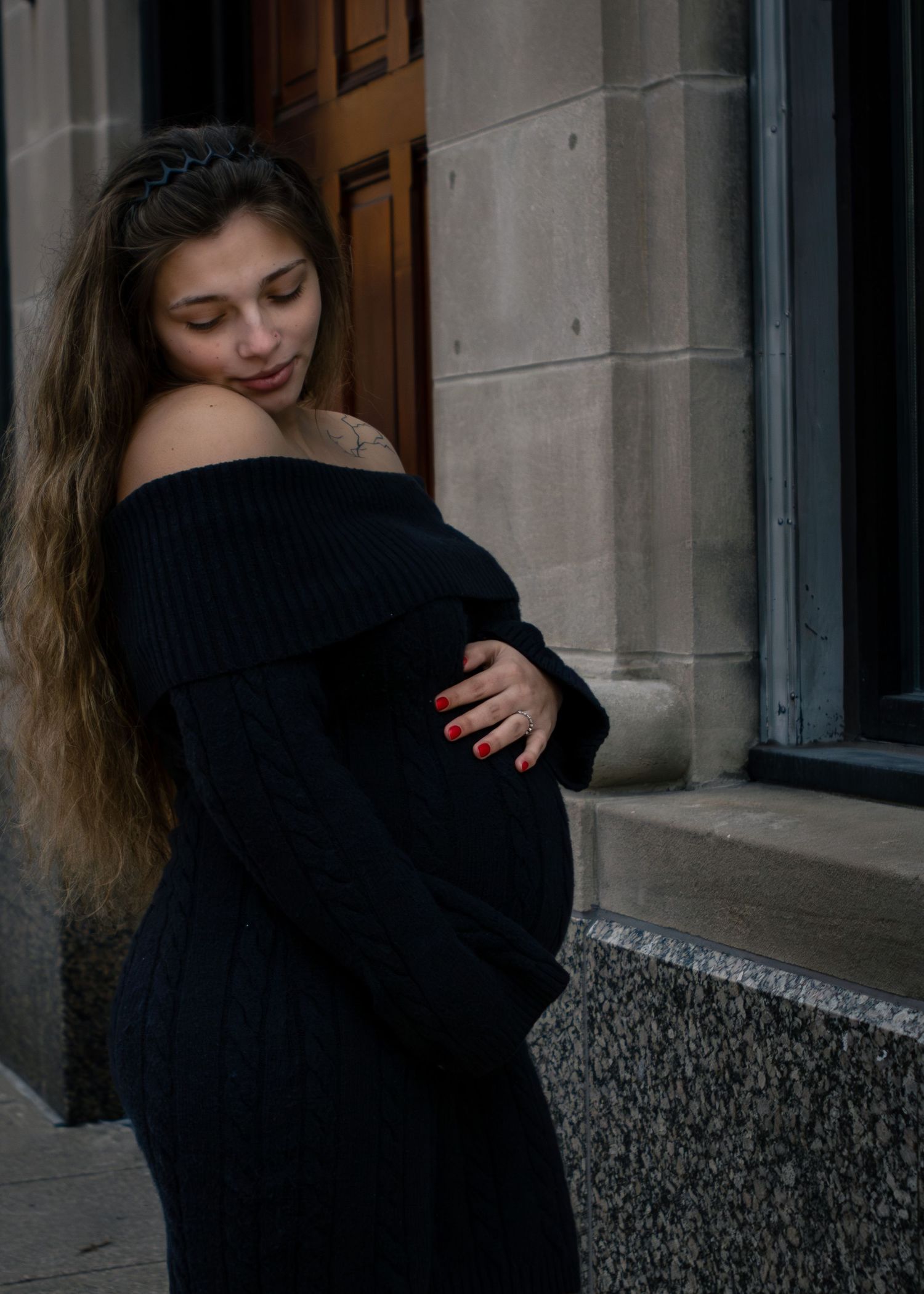 Pregnant woman in black off-shoulder knit dress cradles her baby bump while standing near a stone building entrance.