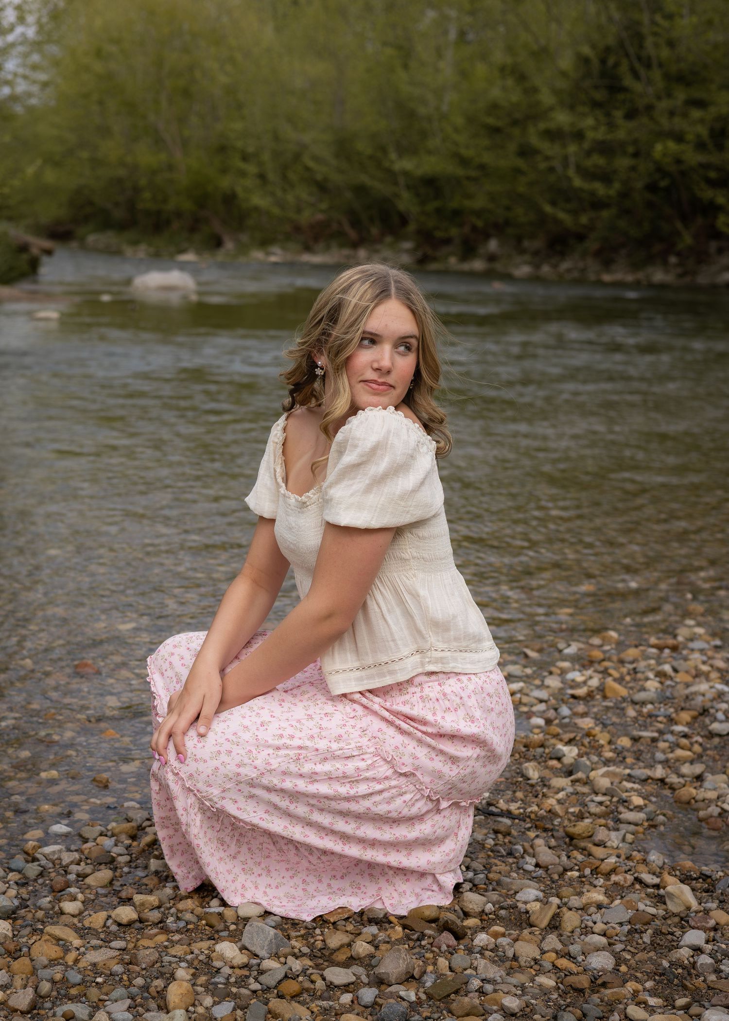 Young woman in cream blouse and pink floral skirt crouching on rocky riverbank with tree-lined stream behind her.