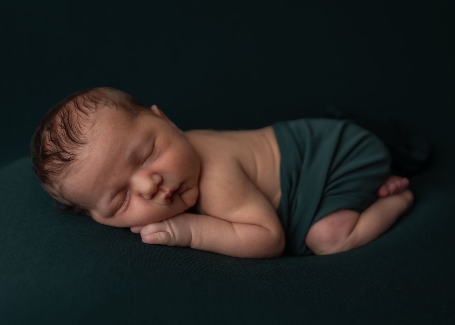 Peaceful sleeping newborn wrapped in teal cloth posed on dark teal background.