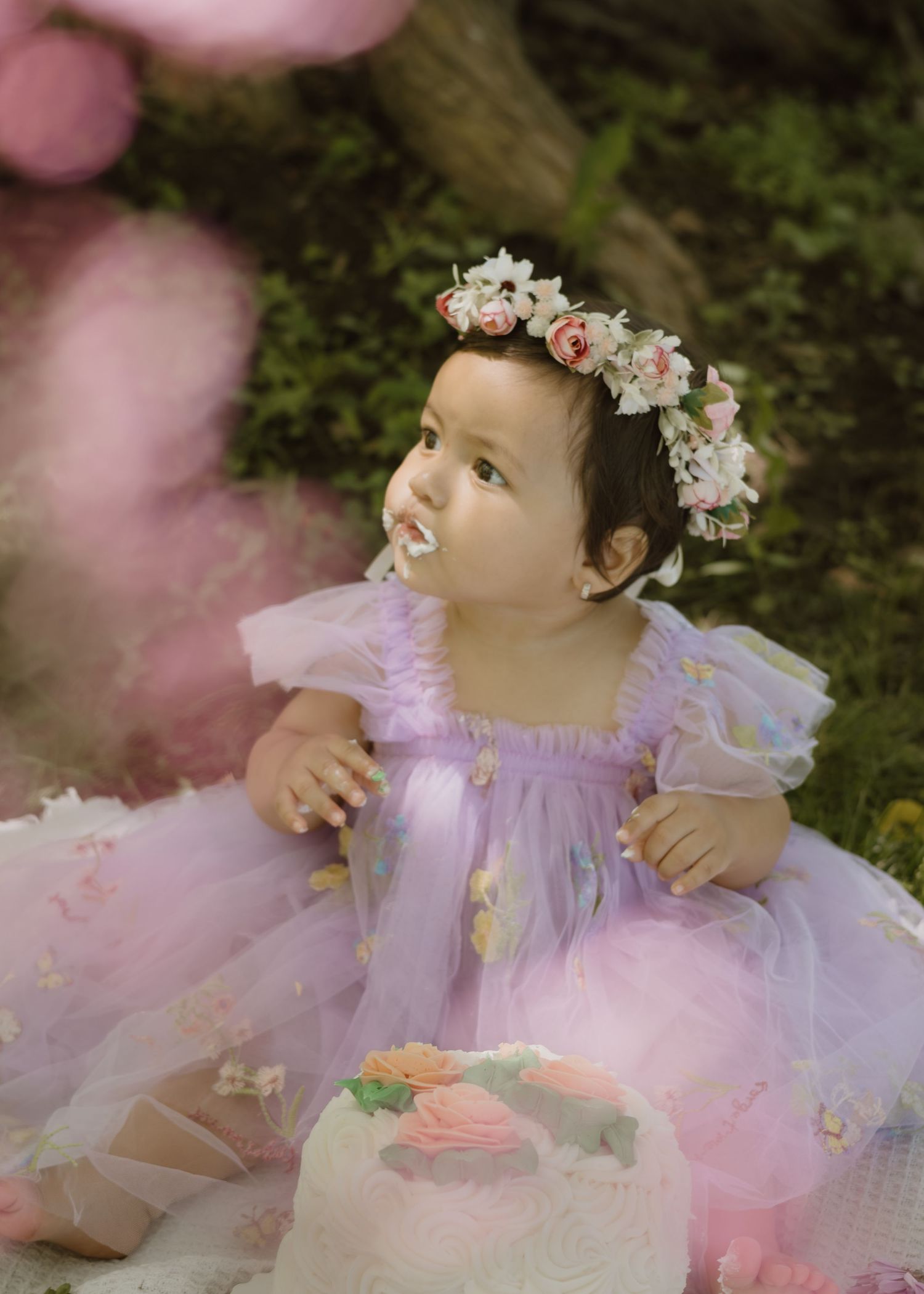 Precious baby girl in purple floral dress and rose crown at outdoor first birthday.