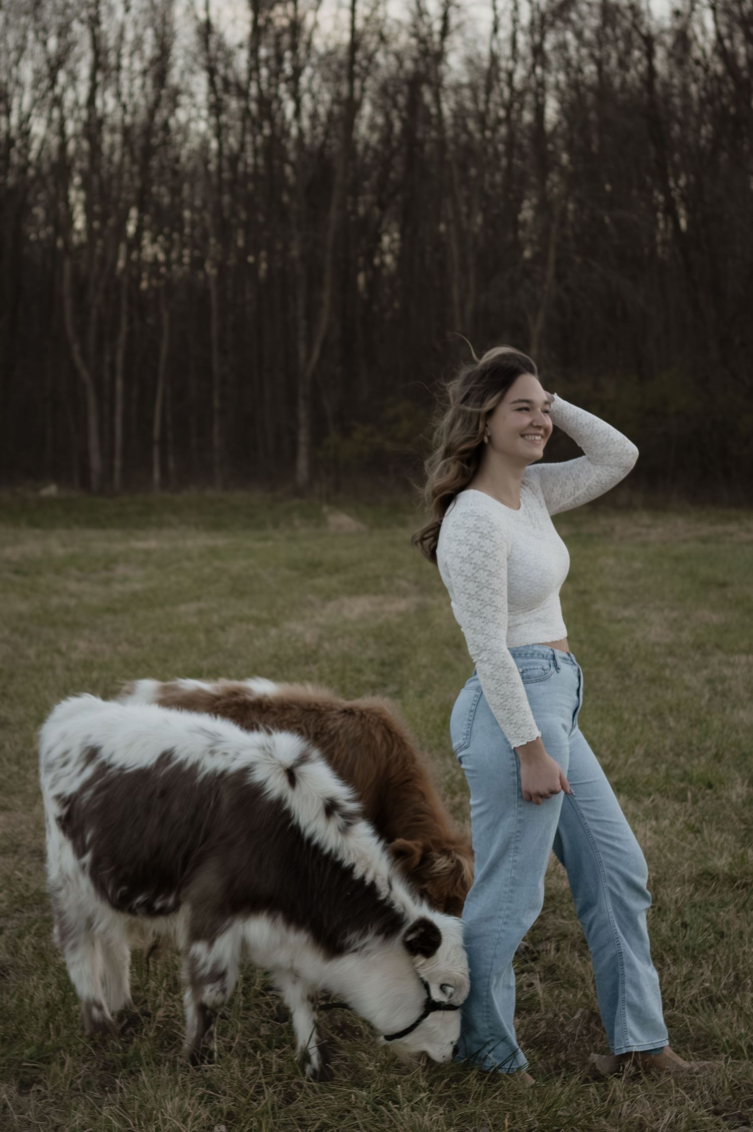 Happy young woman in white top and jeans smiles while standing beside a brown and white miniature cow grazing.