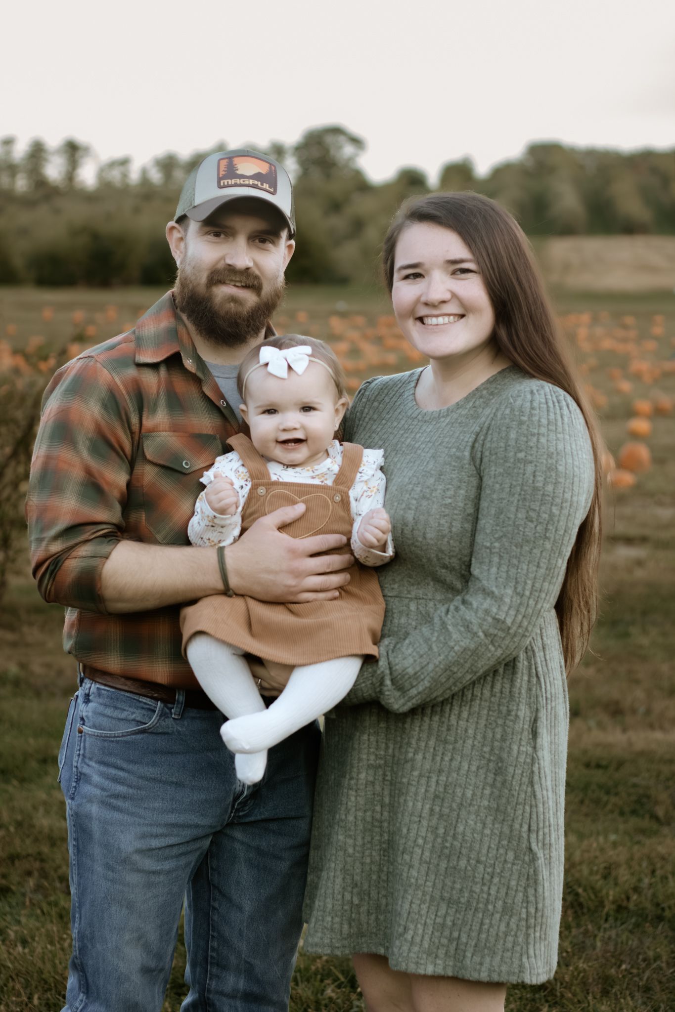 Happy young family with bearded dad, smiling mom, and baby girl posing in a pumpkin patch.