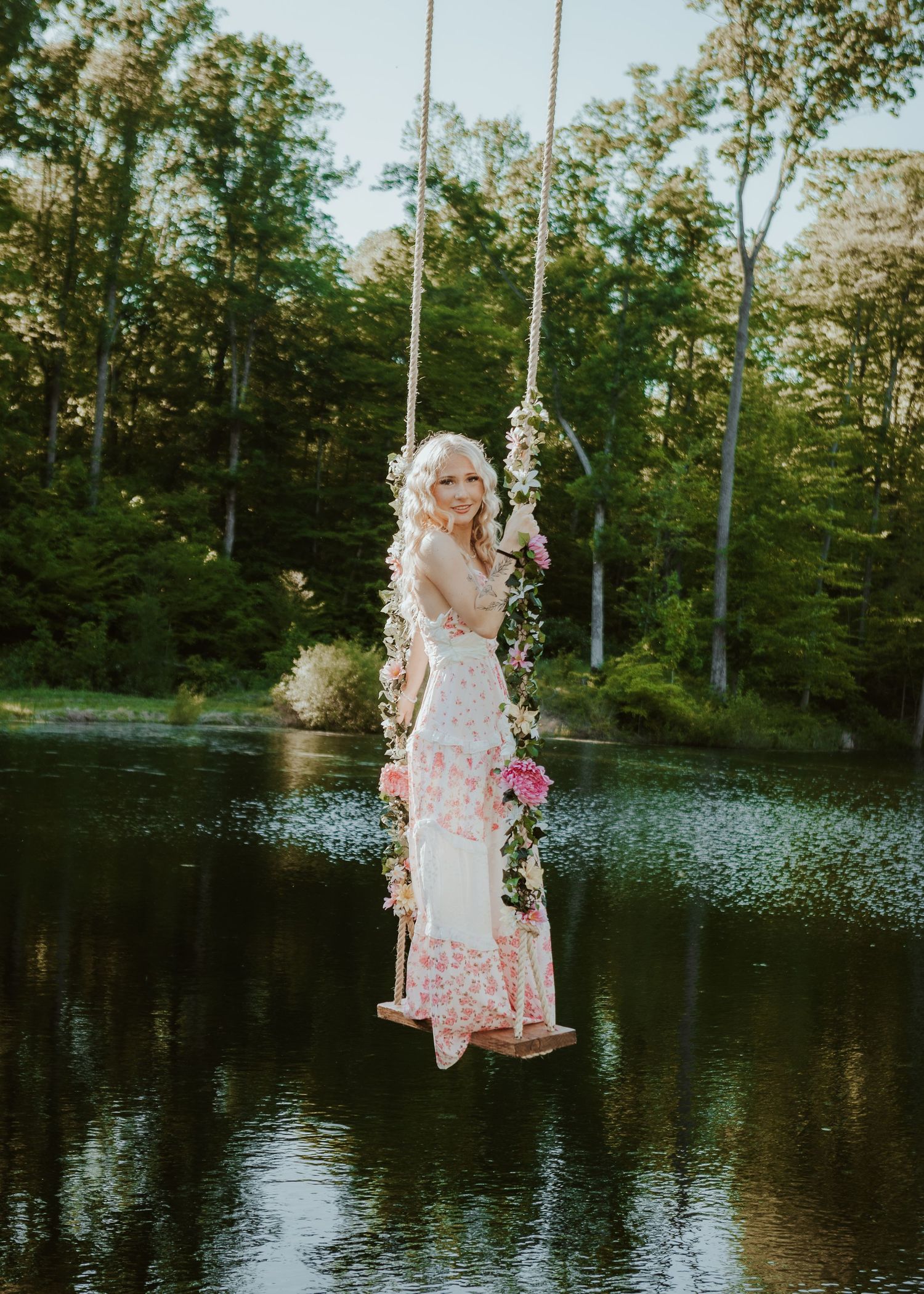 Blonde woman in floral dress sitting on flower-adorned rope swing over a serene lake surrounded by lush green trees.