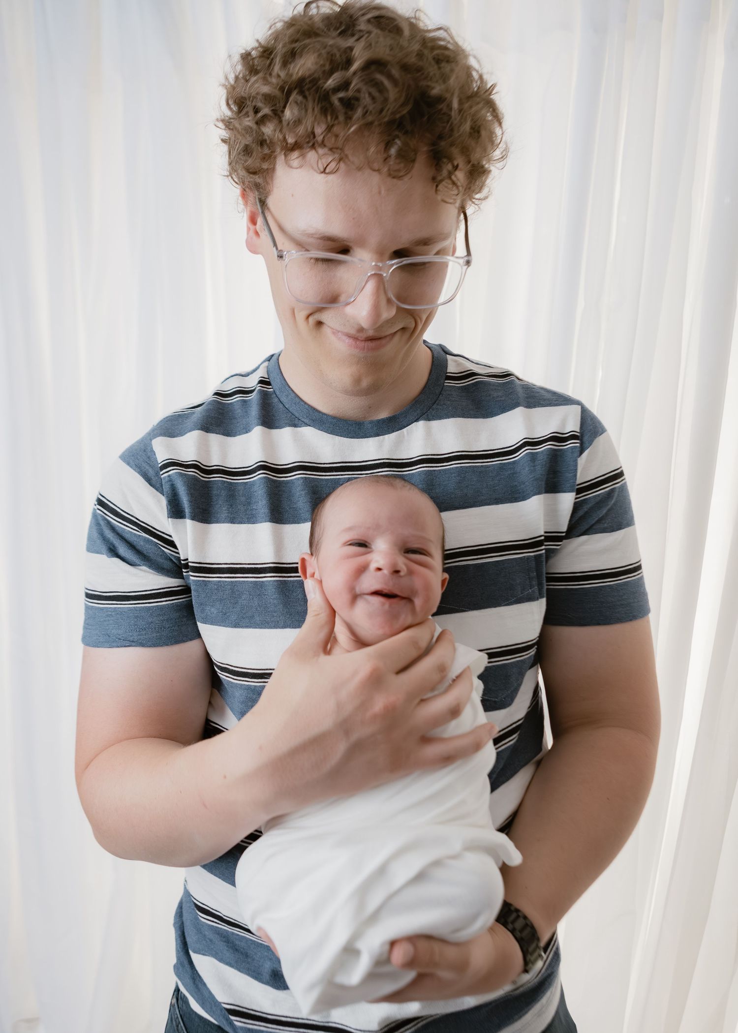 Curly-haired dad with glasses smiles joyfully while holding his swaddled smiling newborn baby.