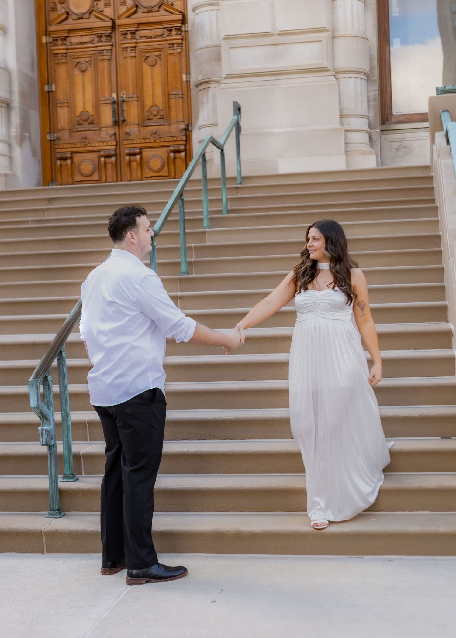 Couple holding hands on grand stone steps outside a historic building in elegant attire.