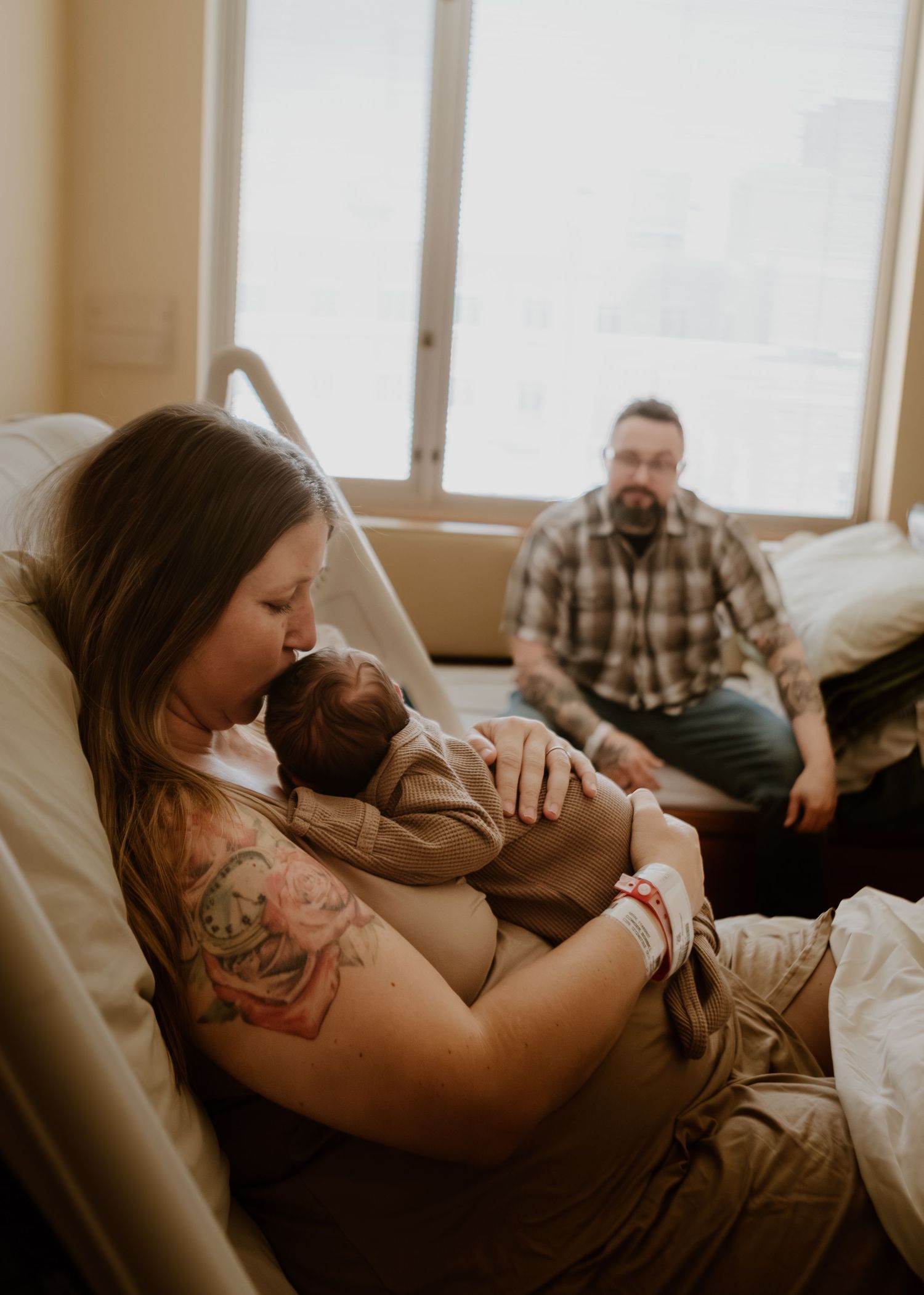 New mother kisses newborn in hospital bed while tattooed father watches lovingly in background.
