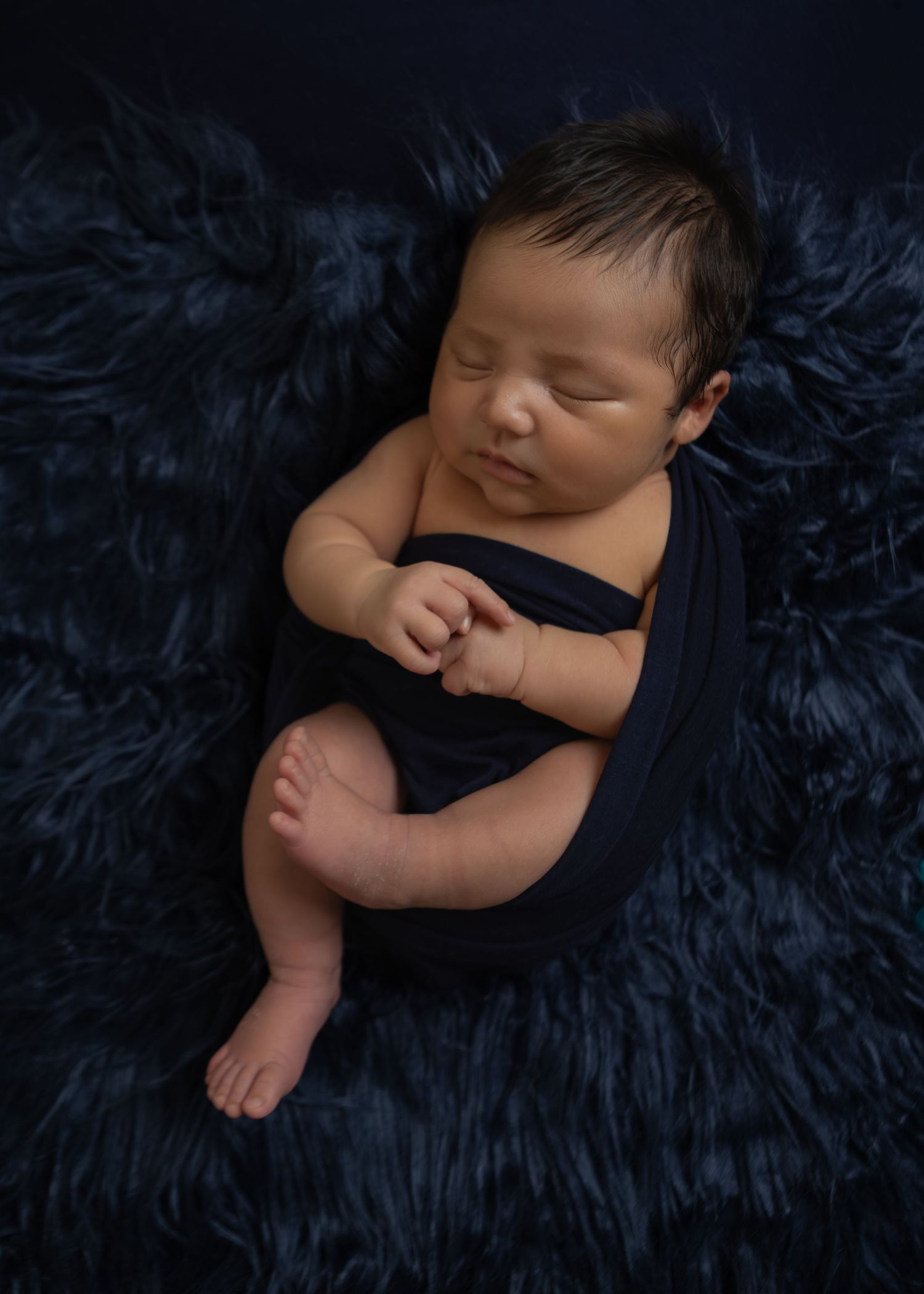 Sleeping newborn wrapped in navy blue swaddle on a dark fur blanket with soft studio lighting.
