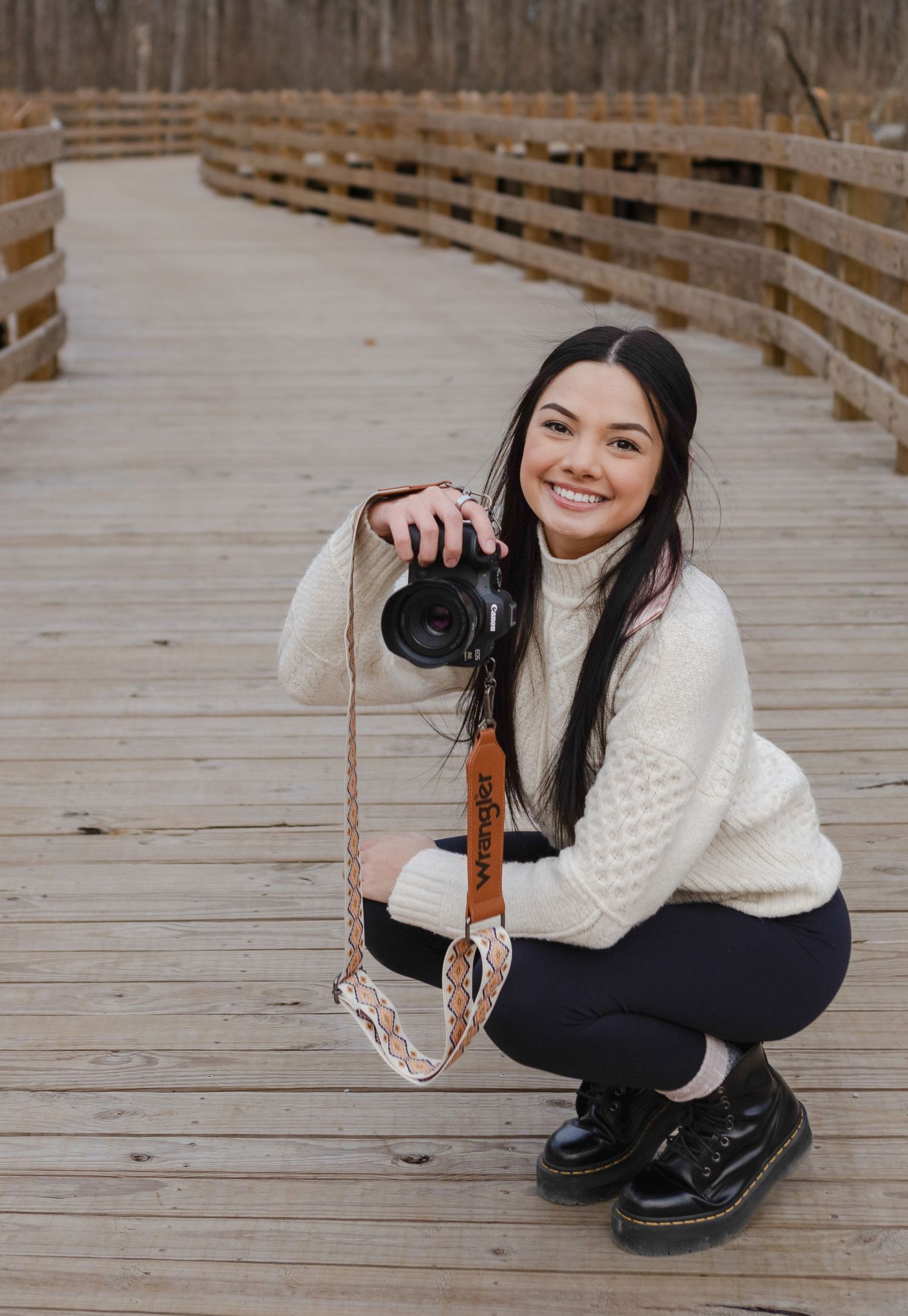 Smiling young woman crouches on wooden bridge holding a DSLR camera with a leather wrist strap.